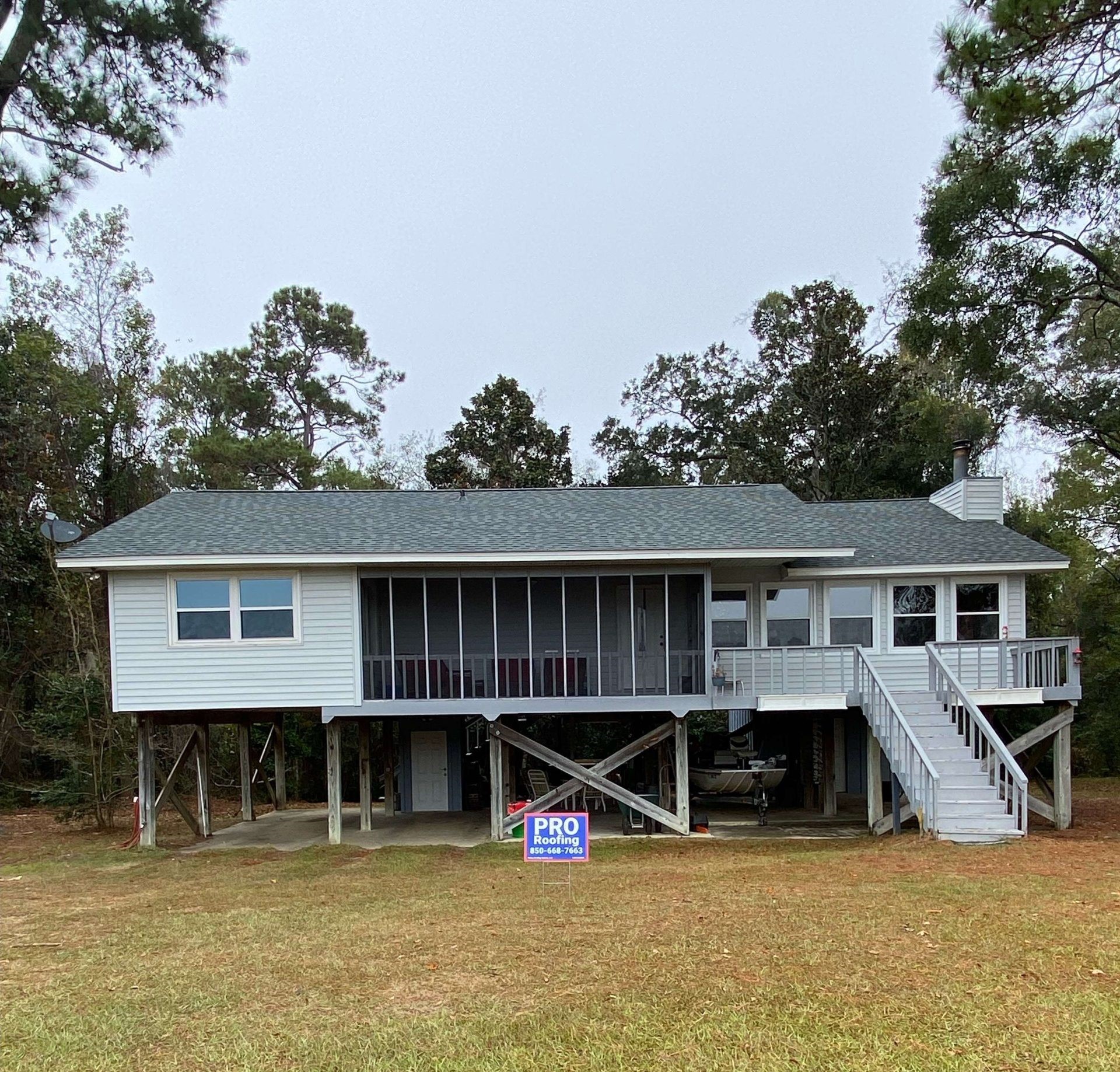 A white elevated house with a screened porch and exterior staircase, surrounded by trees, with a Pro Home Repairs sign on the lawn.