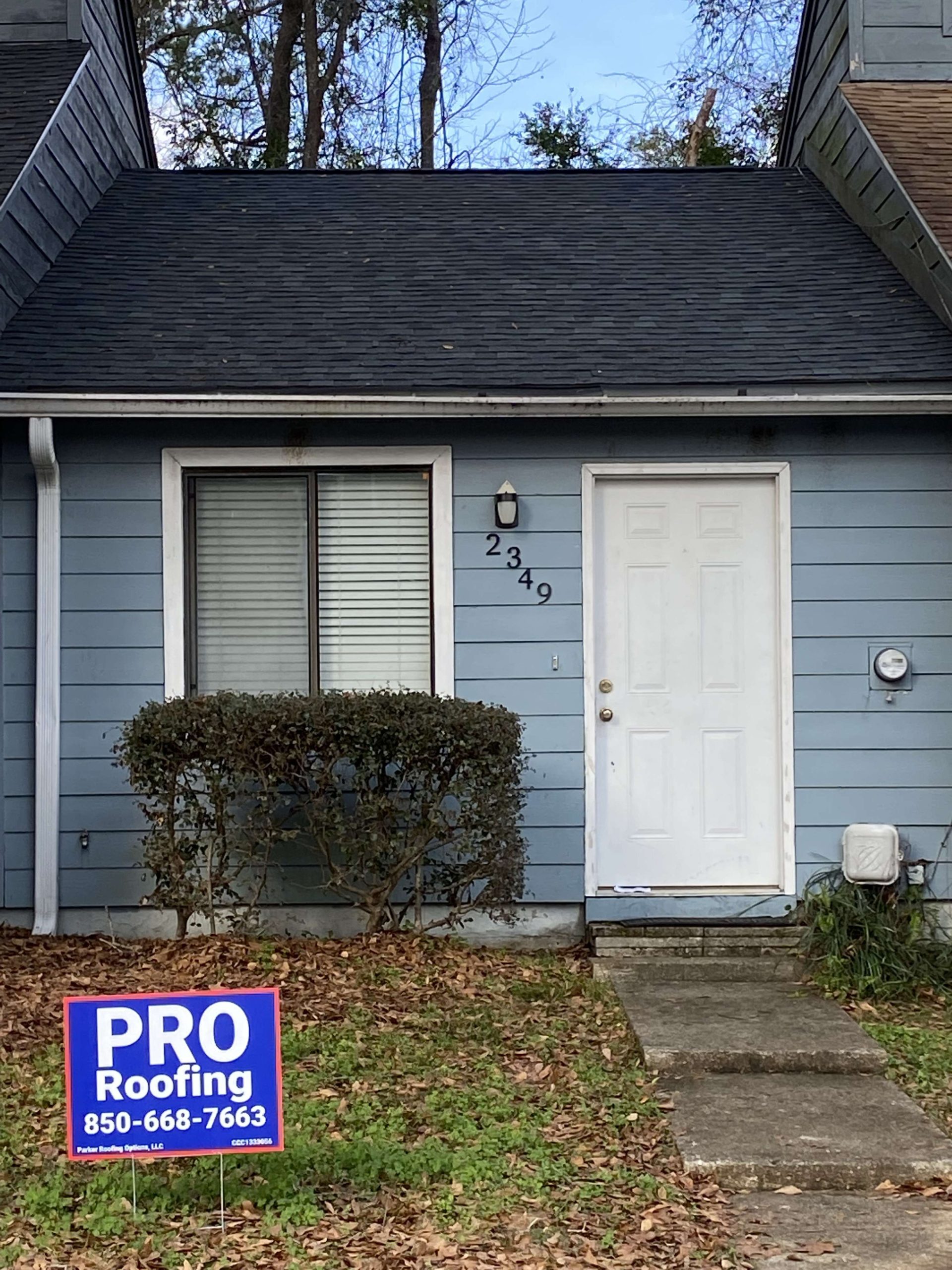 A blue house with a white door and a PRO Roofing sign in the yard, displaying the phone number 850-668-7663. House number 2349 is shown next to the door.