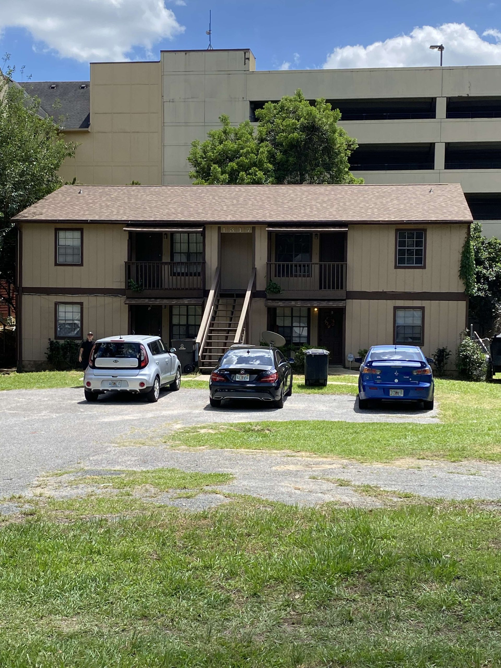 A two-story beige apartment building with an exterior staircase, three parked cars in front, and a parking garage in the background.
