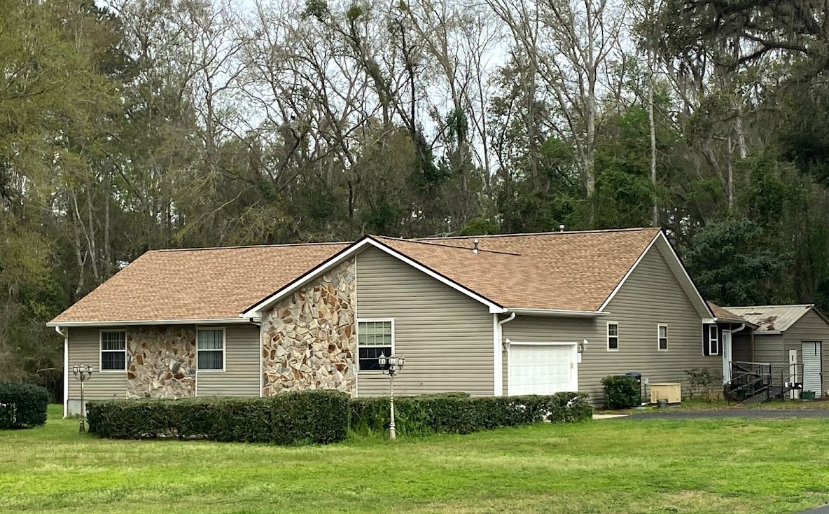A single-story house with tan siding, stone accents, and a brown shingle roof, surrounded by grass and trees in the background.