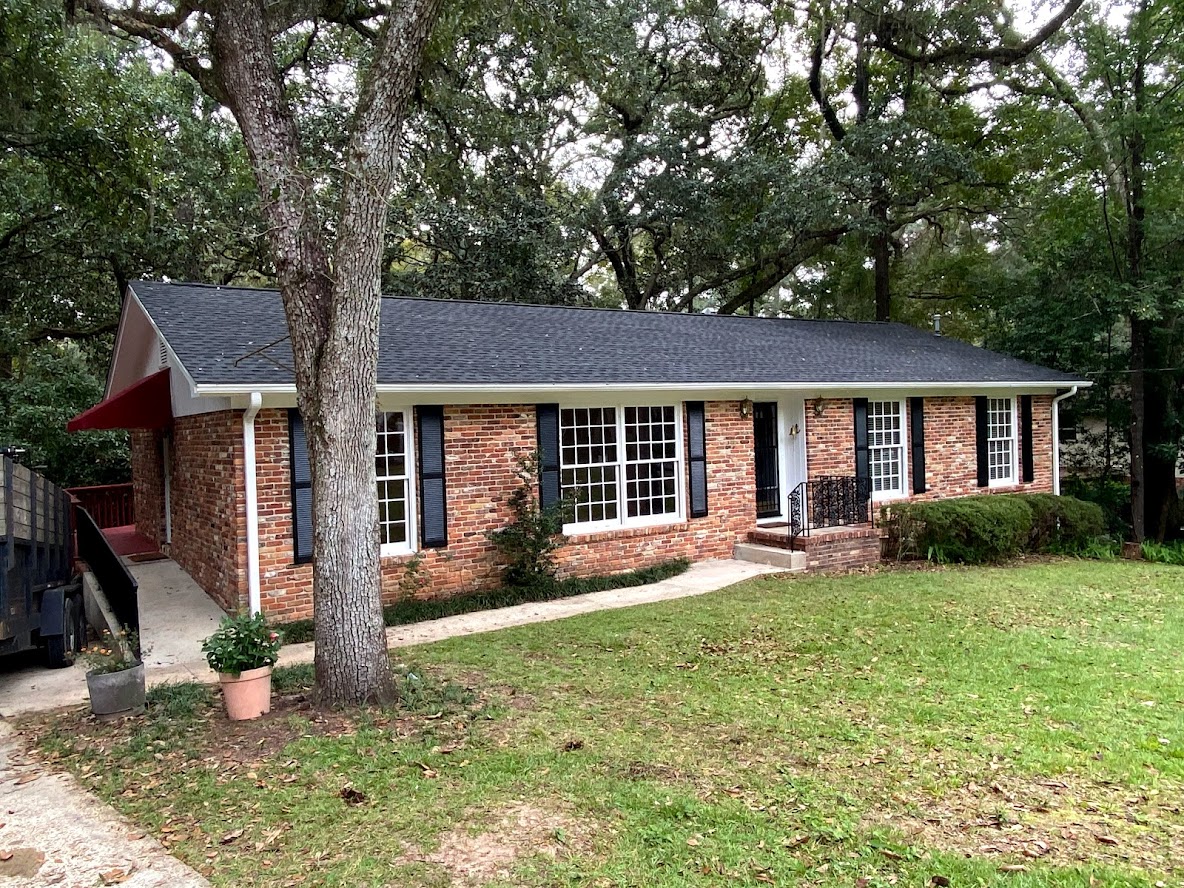 Single-story brick house with black shutters, large windows, a black front door, and a small front porch, surrounded by trees and grass.