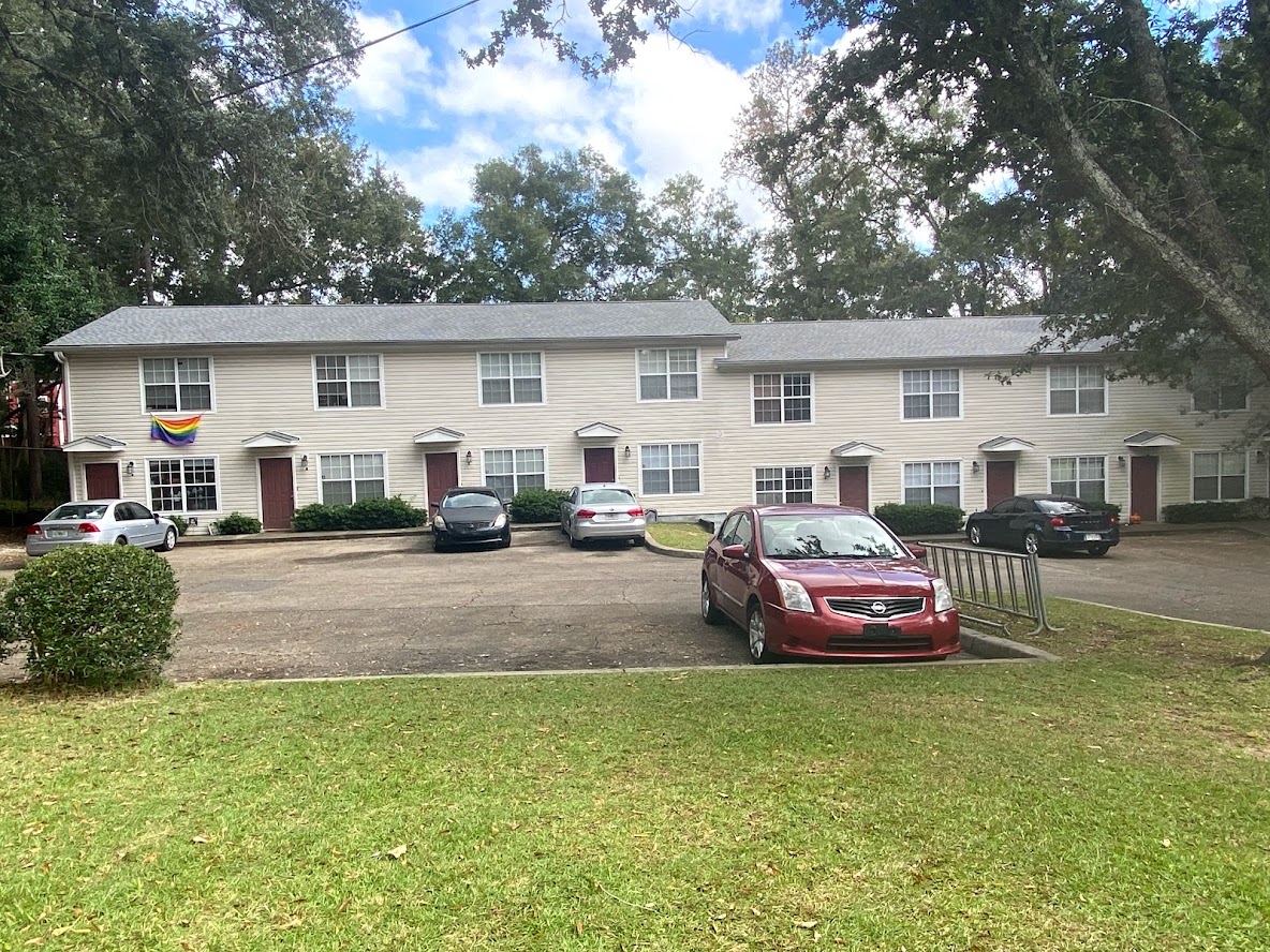 A two-story apartment building with white siding and red doors, several parked cars in front, and a rainbow flag hanging by one entrance.