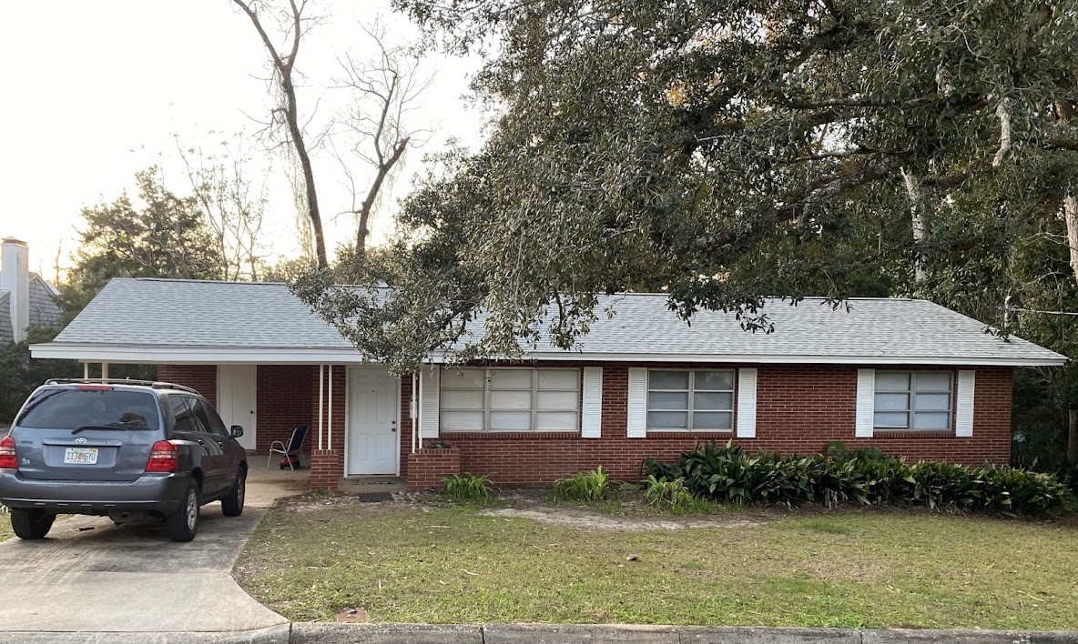 A single-story red brick house with a gray roof, a driveway with a parked SUV, a small front porch, and a patchy lawn, surrounded by trees.