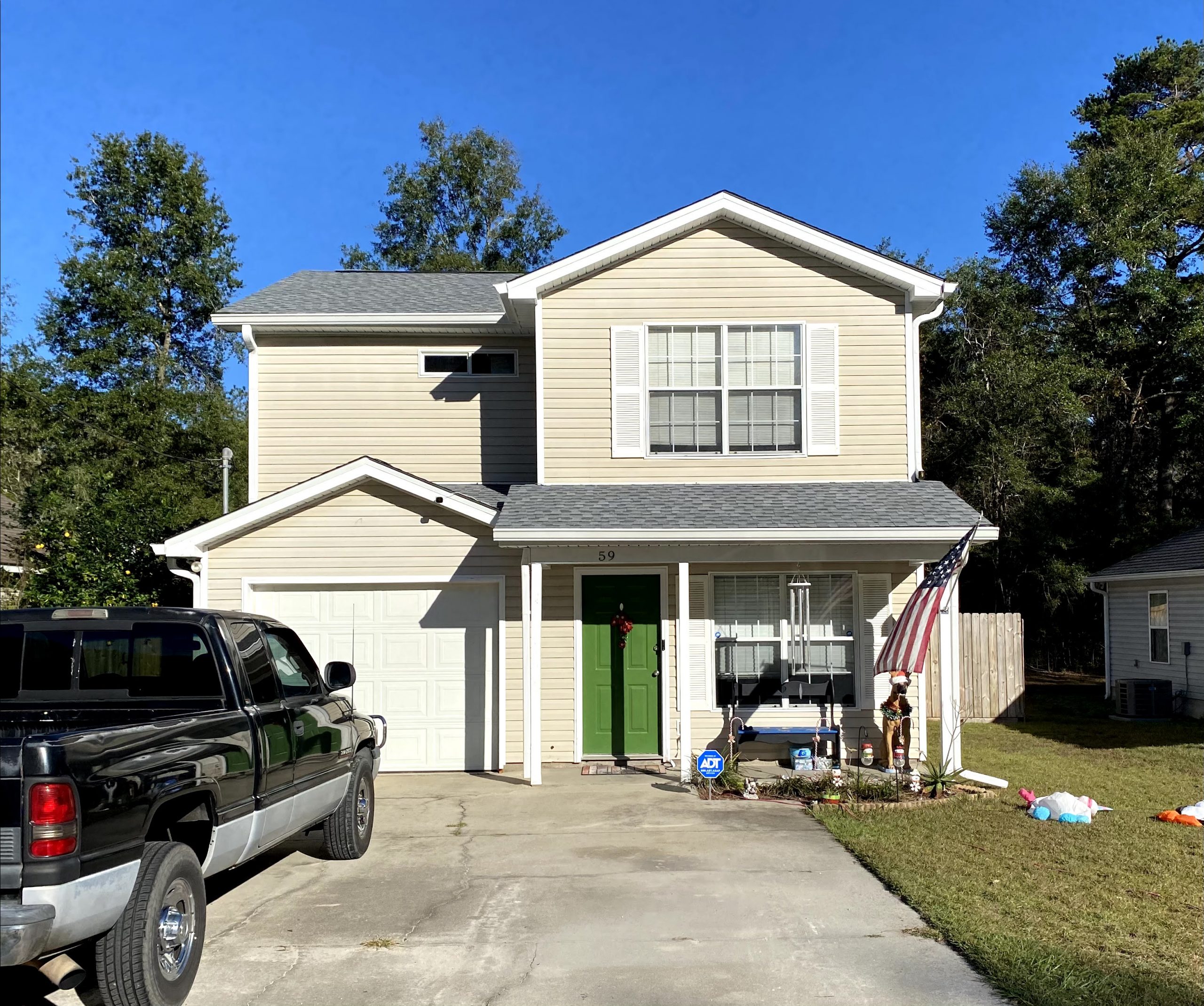 Two-story beige house with green front door, single-car garage, and an American flag by the entrance. A black truck is parked in the driveway on a clear, sunny day.