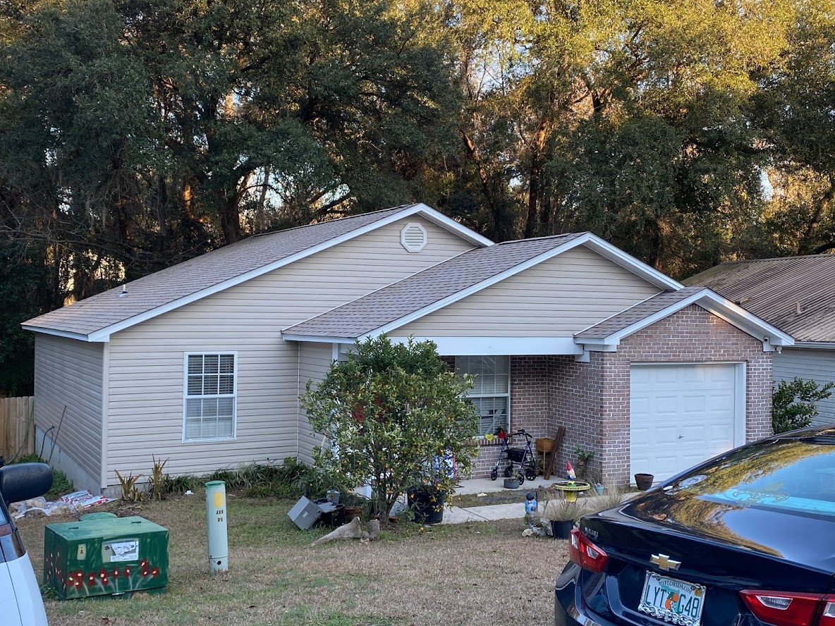 Single-story house with beige siding, a front porch, and attached garage. Items are scattered near the porch, with a black car and green utility box in the driveway. Trees line the background.