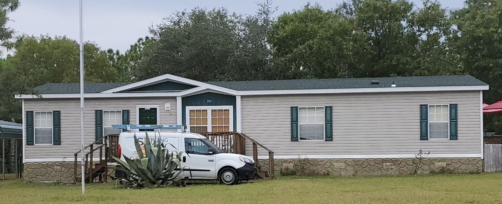 A white single-story manufactured home with green shutters, a wooden porch, and a white work van parked in front; a large plant is near the steps.