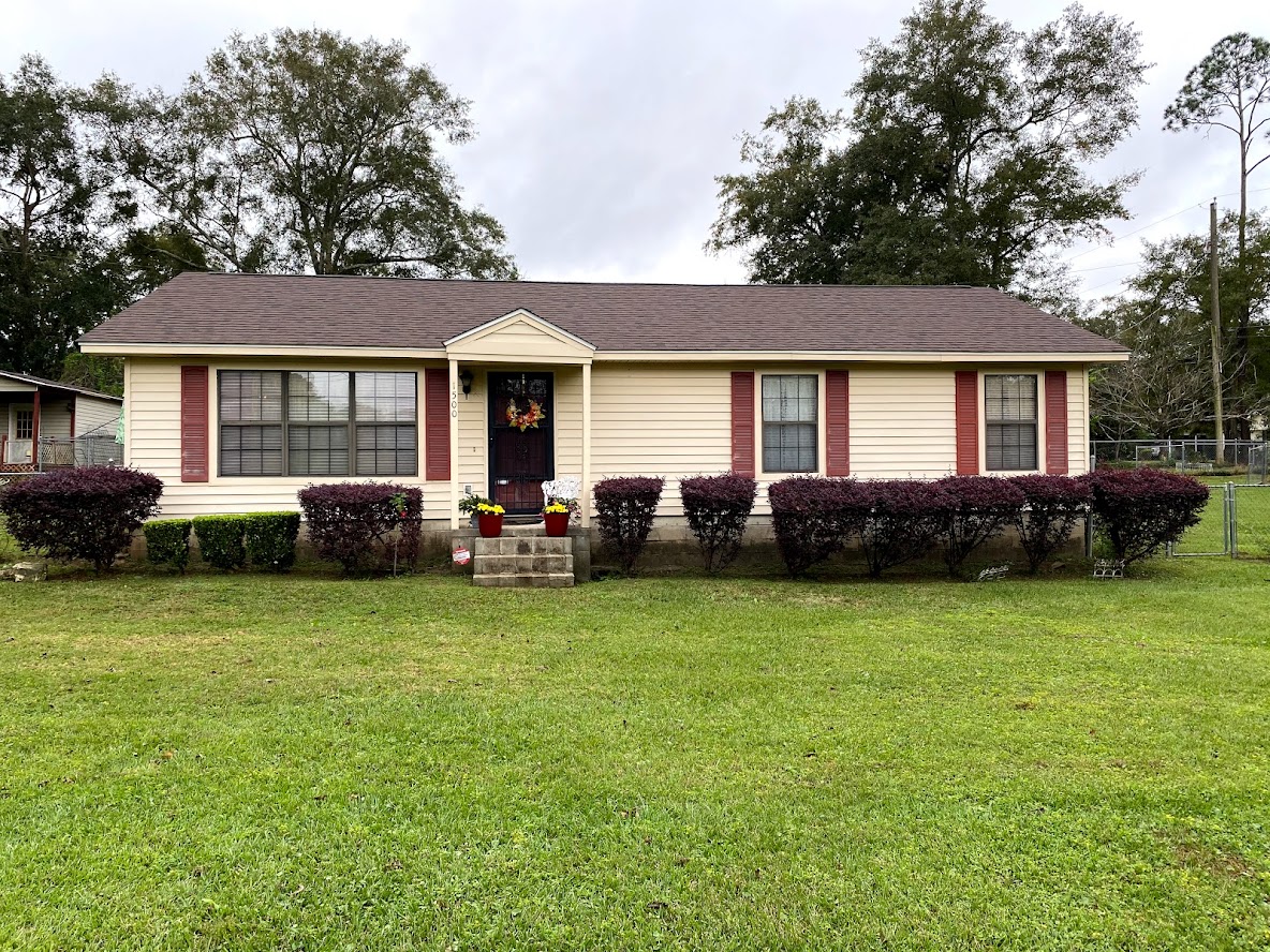Single-story beige house with red shutters, front steps, a dark front door with a wreath, trimmed hedges, and a well-kept lawn. Trees and a fence are visible in the background.