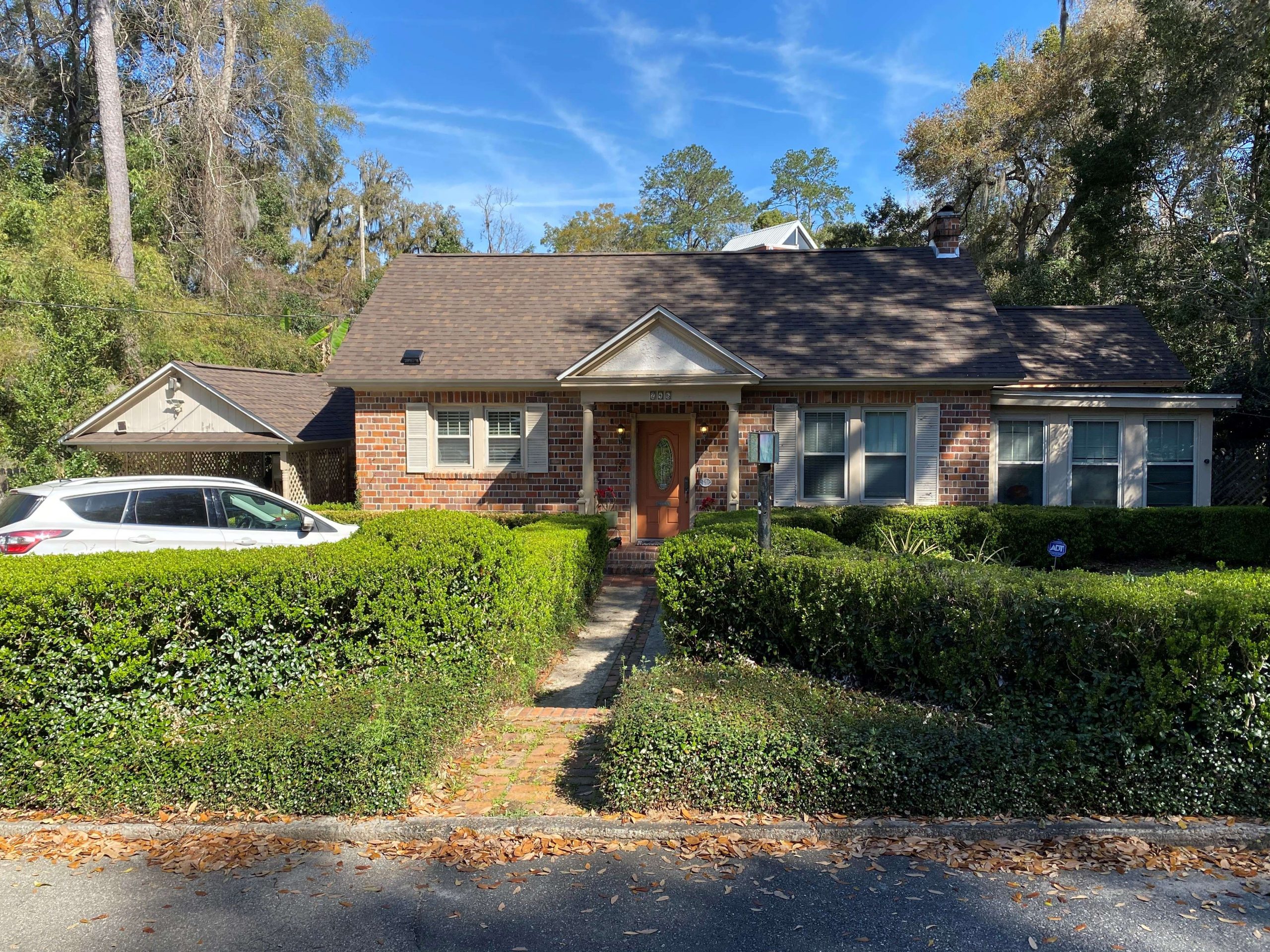 A single-story brick house with a brown roof, front porch, and trimmed hedges, with a white SUV parked in the driveway on a sunny day.