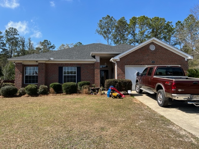 A single-story brick house with a front lawn, shrubs, and a driveway where a red pickup truck and childrens toys are visible. Pine trees are in the background under a blue sky.