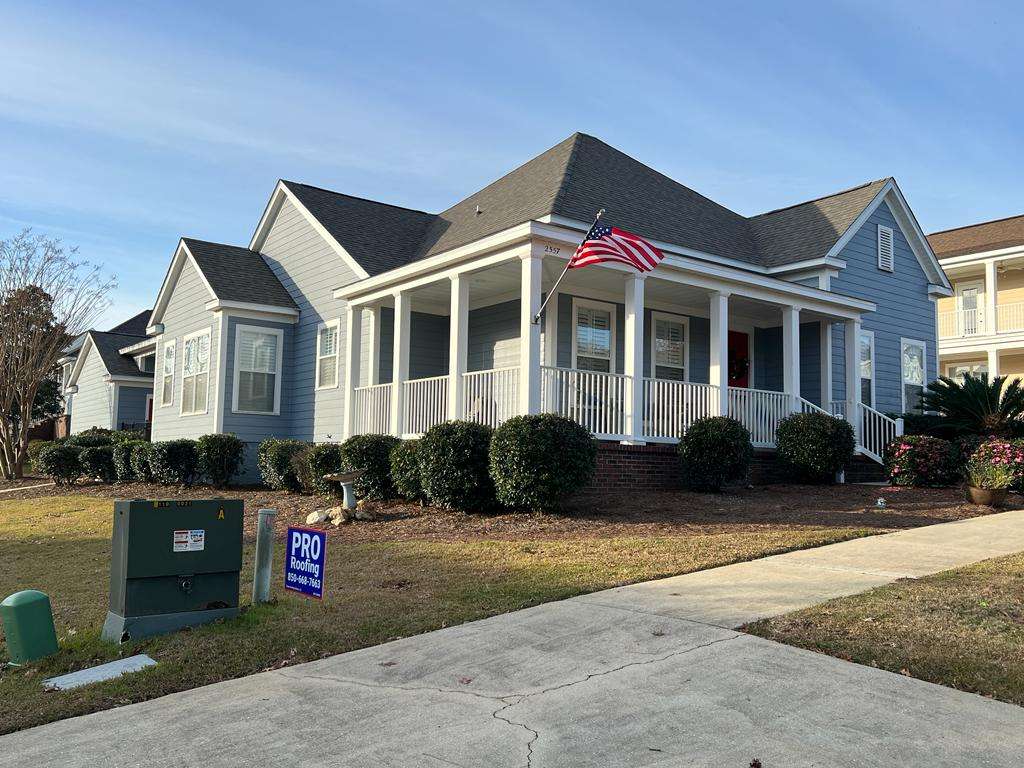 A blue single-story house with a front porch, an American flag, manicured bushes, and a Pro Roofing sign in the yard.