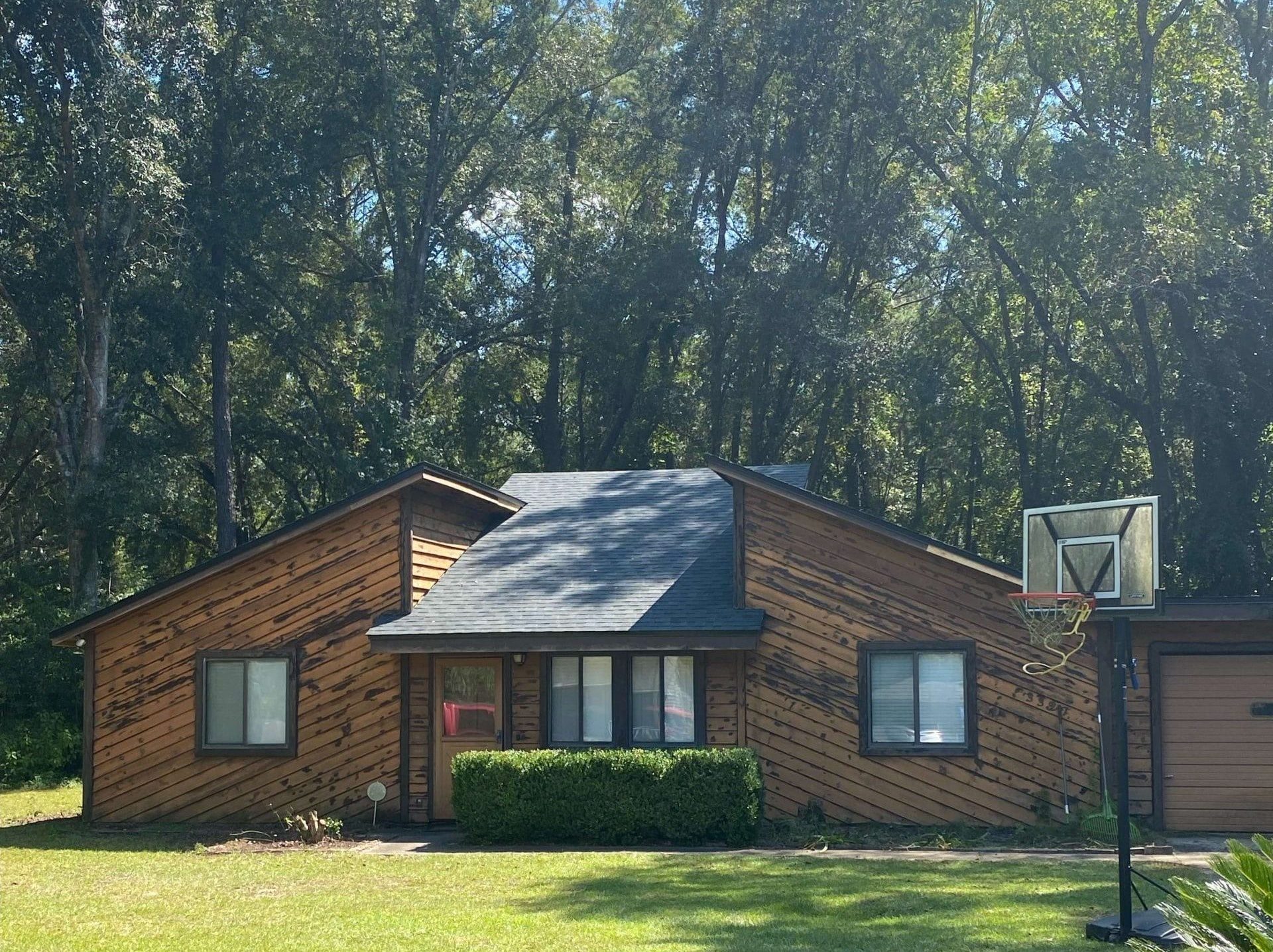 Single-story wooden house with diagonal siding, black roof, bushes in front, and a basketball hoop in the driveway, set against a backdrop of tall trees.