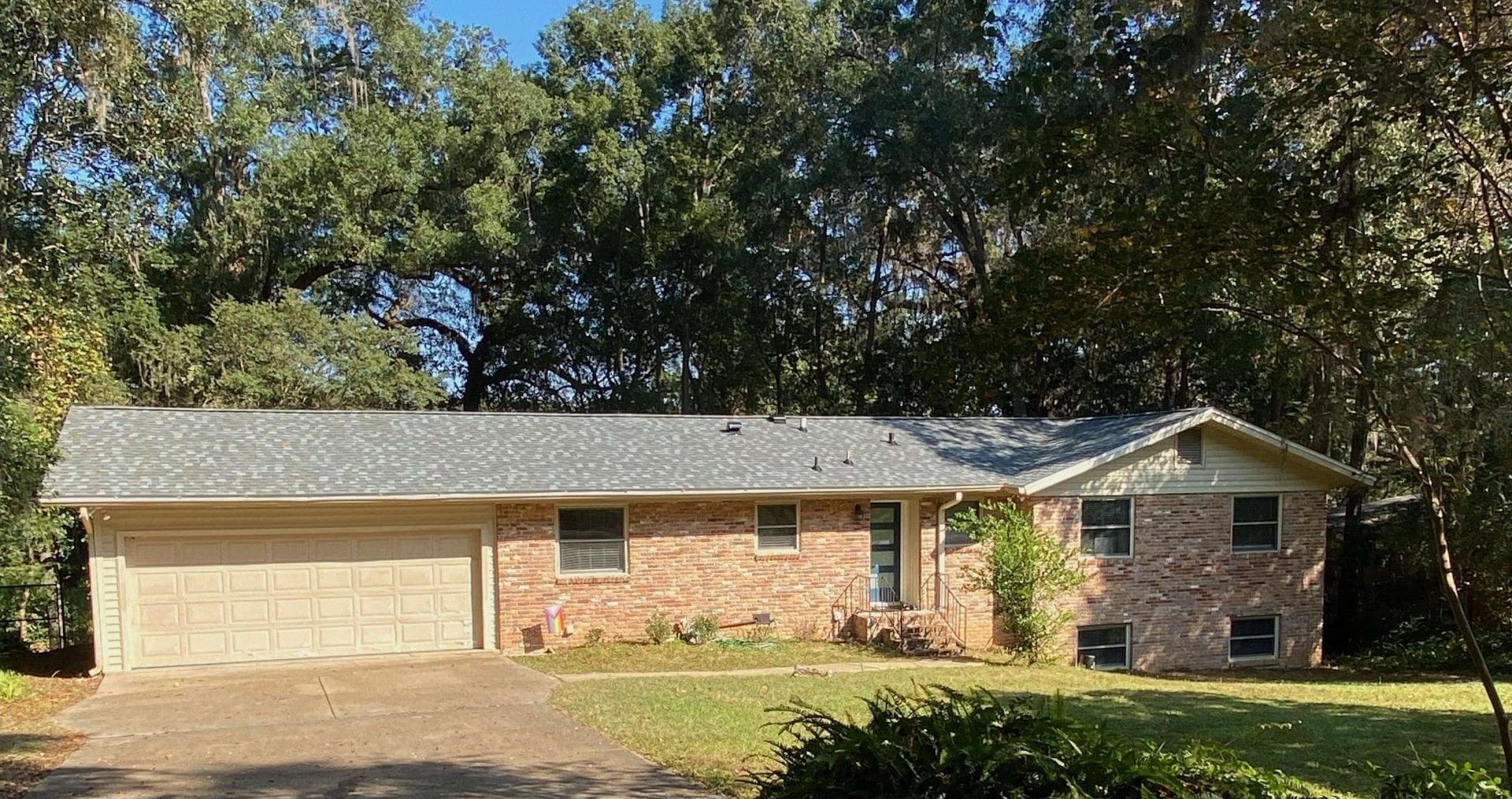 Single-story brick house with attached two-car garage, surrounded by trees and a grassy lawn, under a clear blue sky.