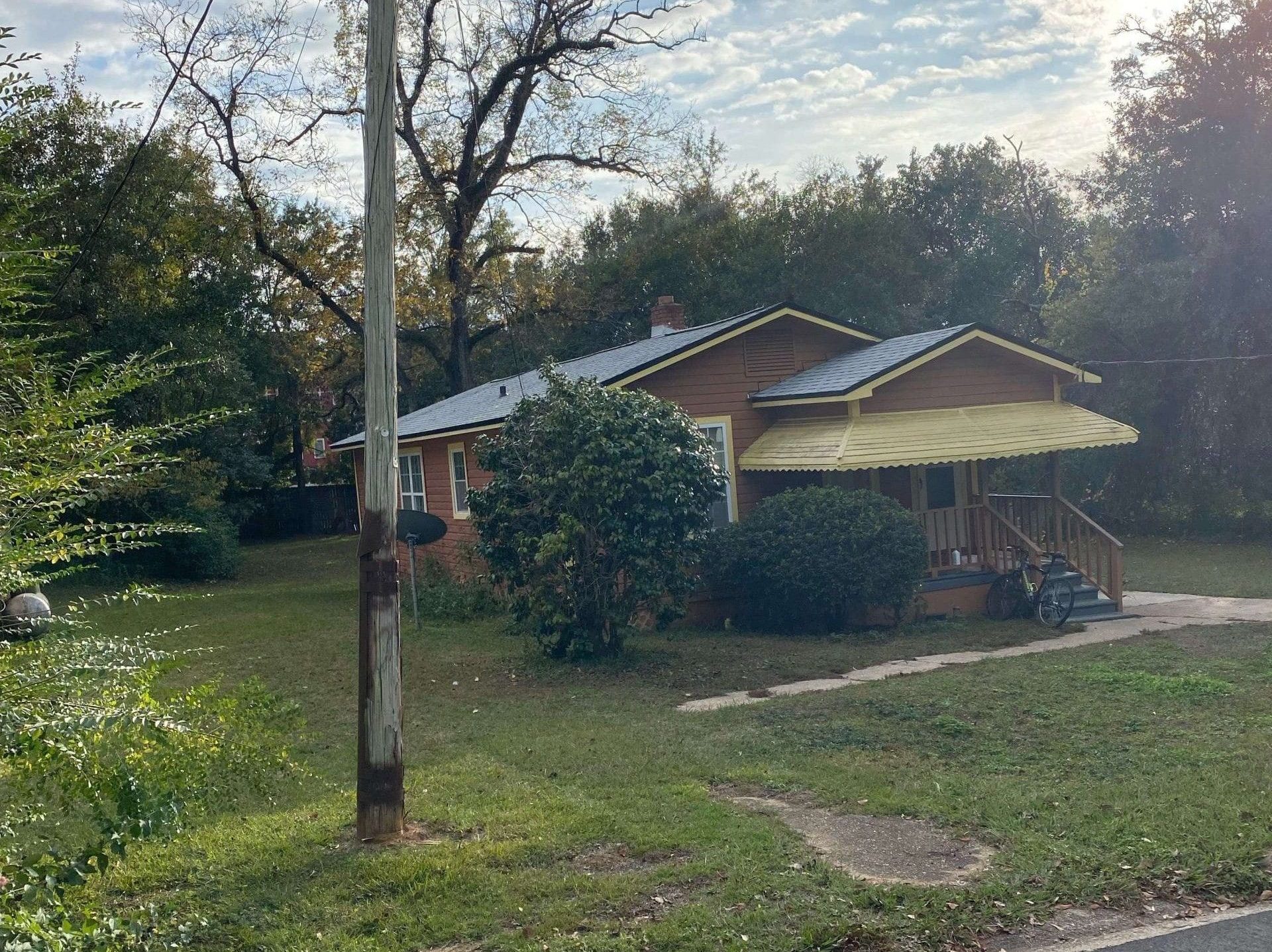 A single-story house with a yellow awning, front porch, and bicycle parked outside, surrounded by trees and grass under a partly cloudy sky.
