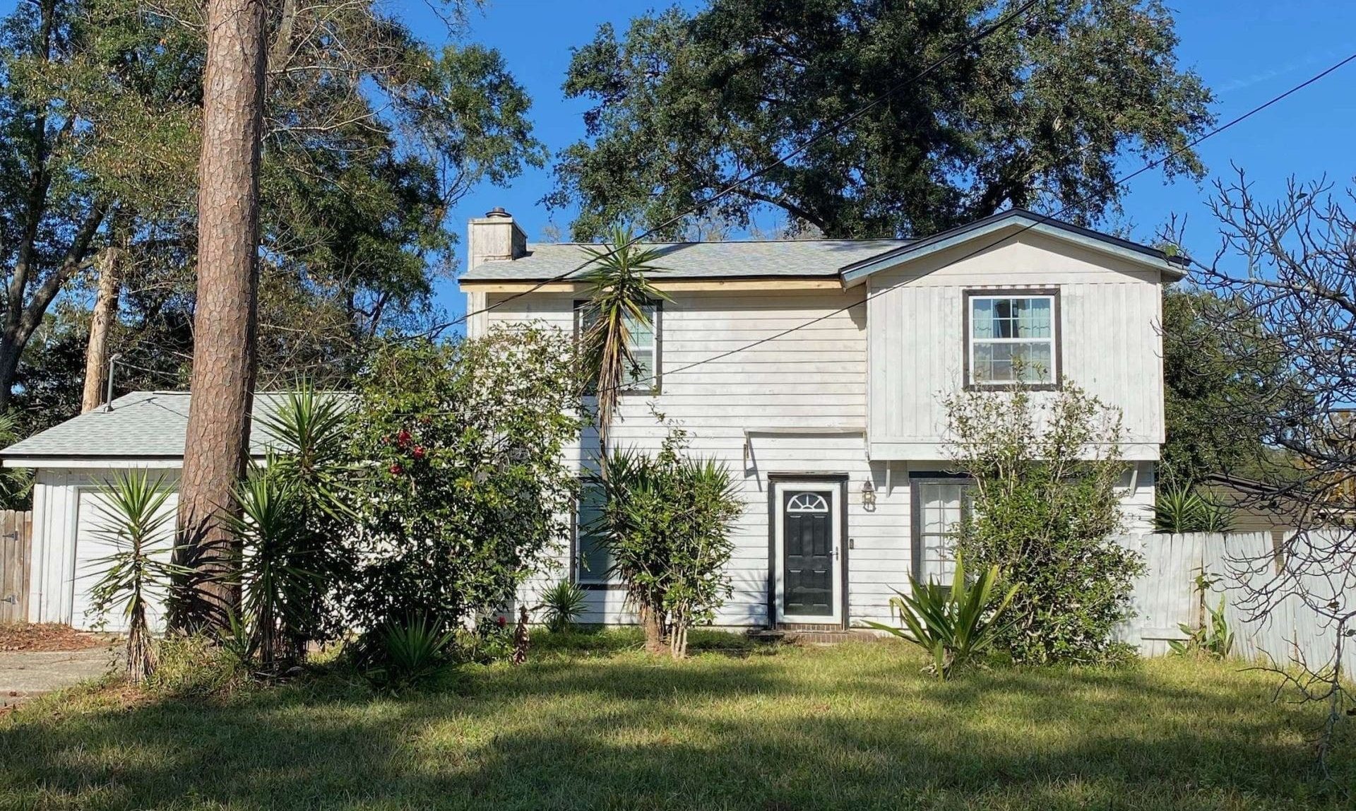 Two-story white house with a black front door, surrounded by overgrown bushes and palm trees, under a clear blue sky.