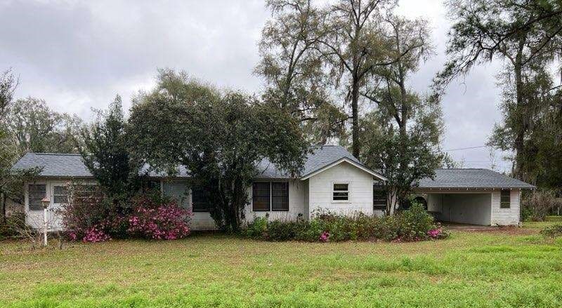 Single-story white house with dark roof, overgrown bushes, pink flowers in front, large grassy yard, and an attached carport on the right side under cloudy skies.