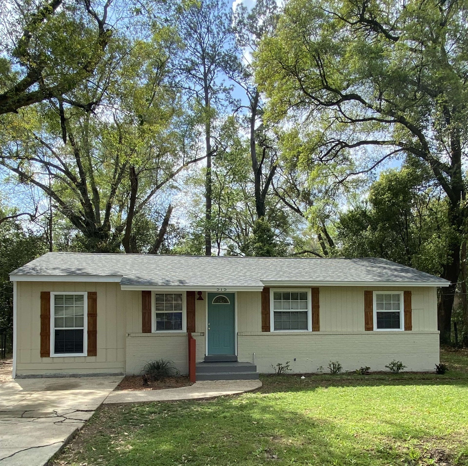 A small, single-story house with tan siding, wooden shutters, a teal front door, and a gray roof, surrounded by trees and a lawn.