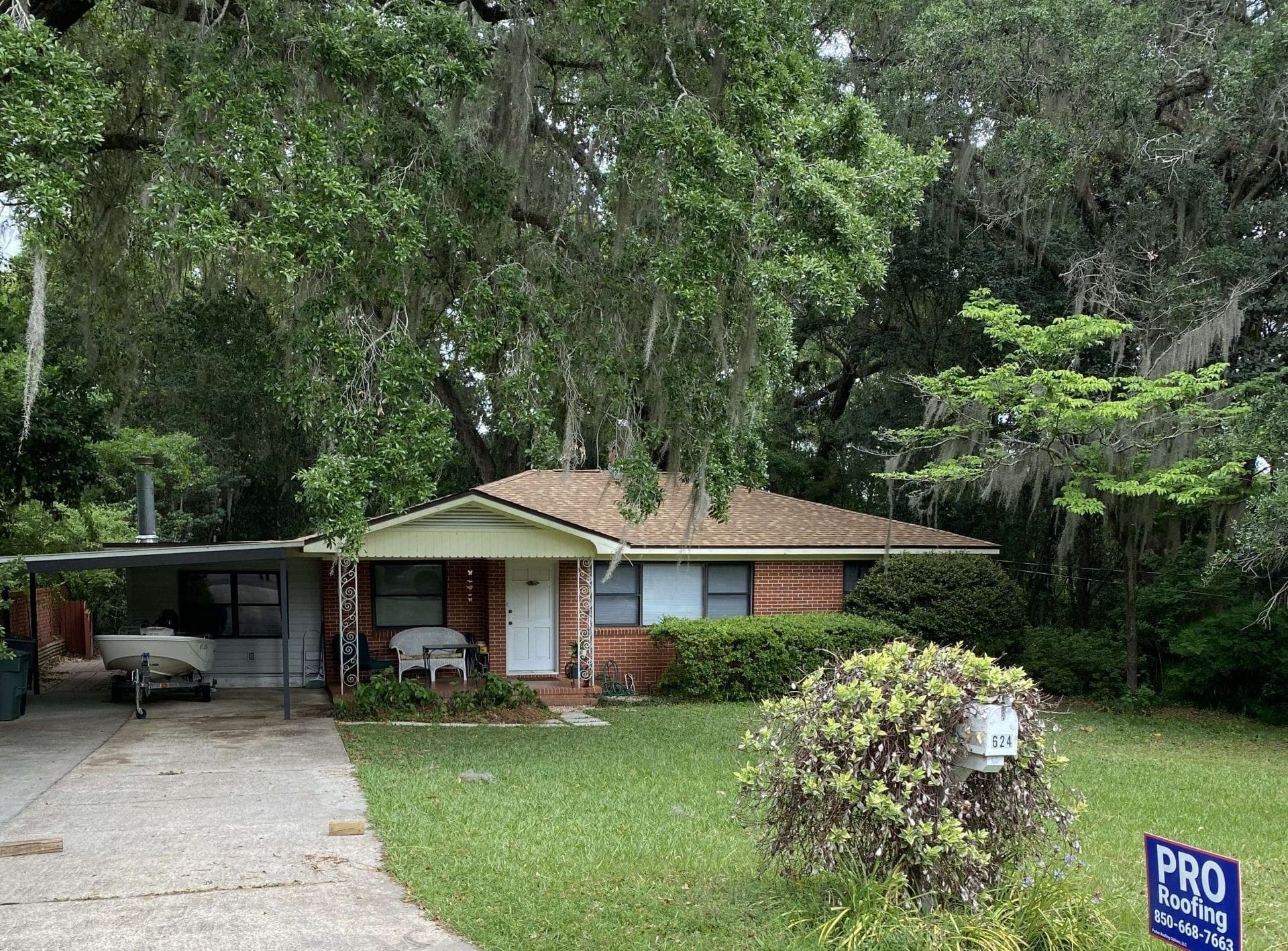 A one-story brick house with a carport, boat, and lawn, surrounded by large trees. A PRO Roofing sign is in the grass near the mailbox.