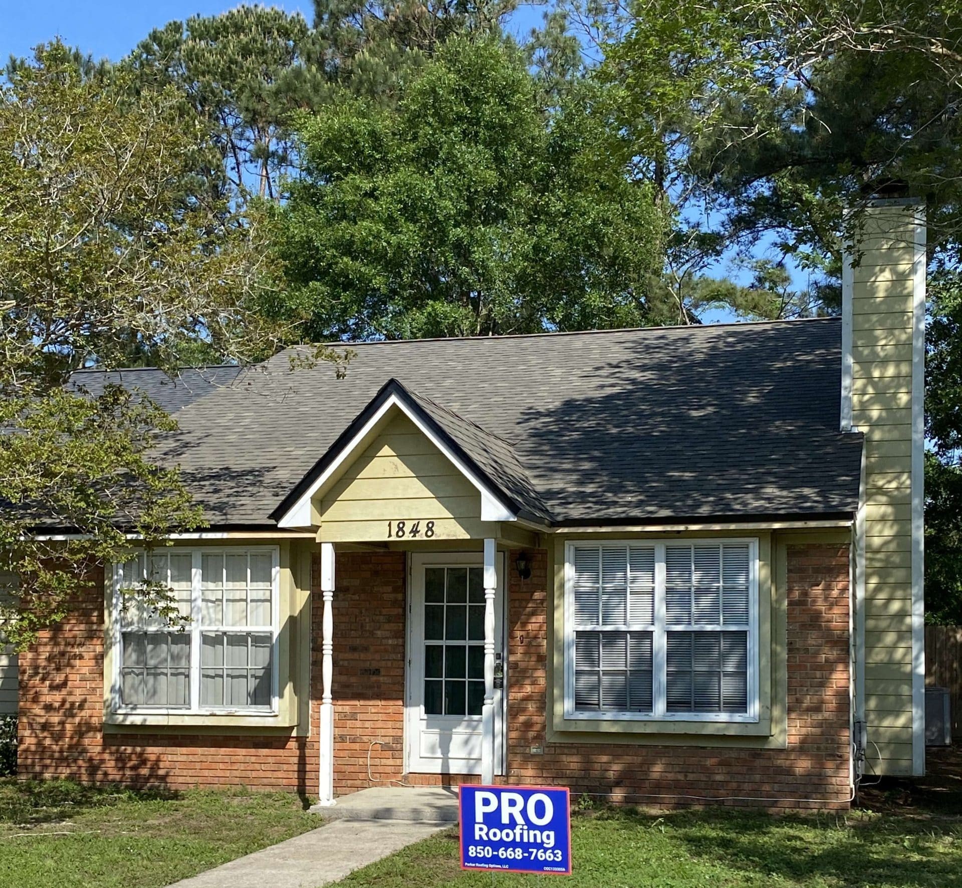 Single-story brick house with yellow trim, a central door, two front windows, and a PRO Roofing sign with a phone number in the yard.