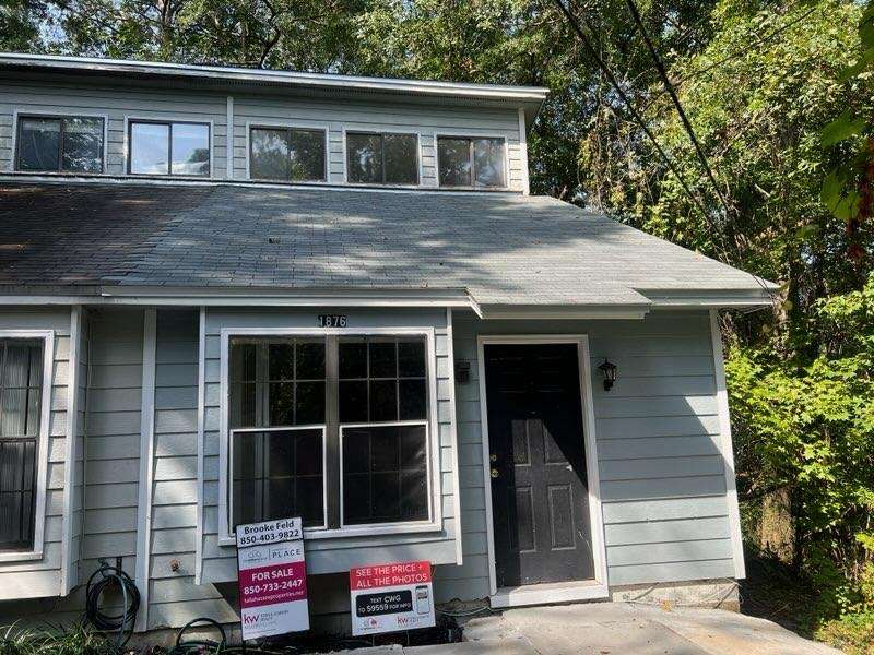 A two-story light blue house with a black front door and a For Sale sign posted in front, surrounded by trees and greenery.