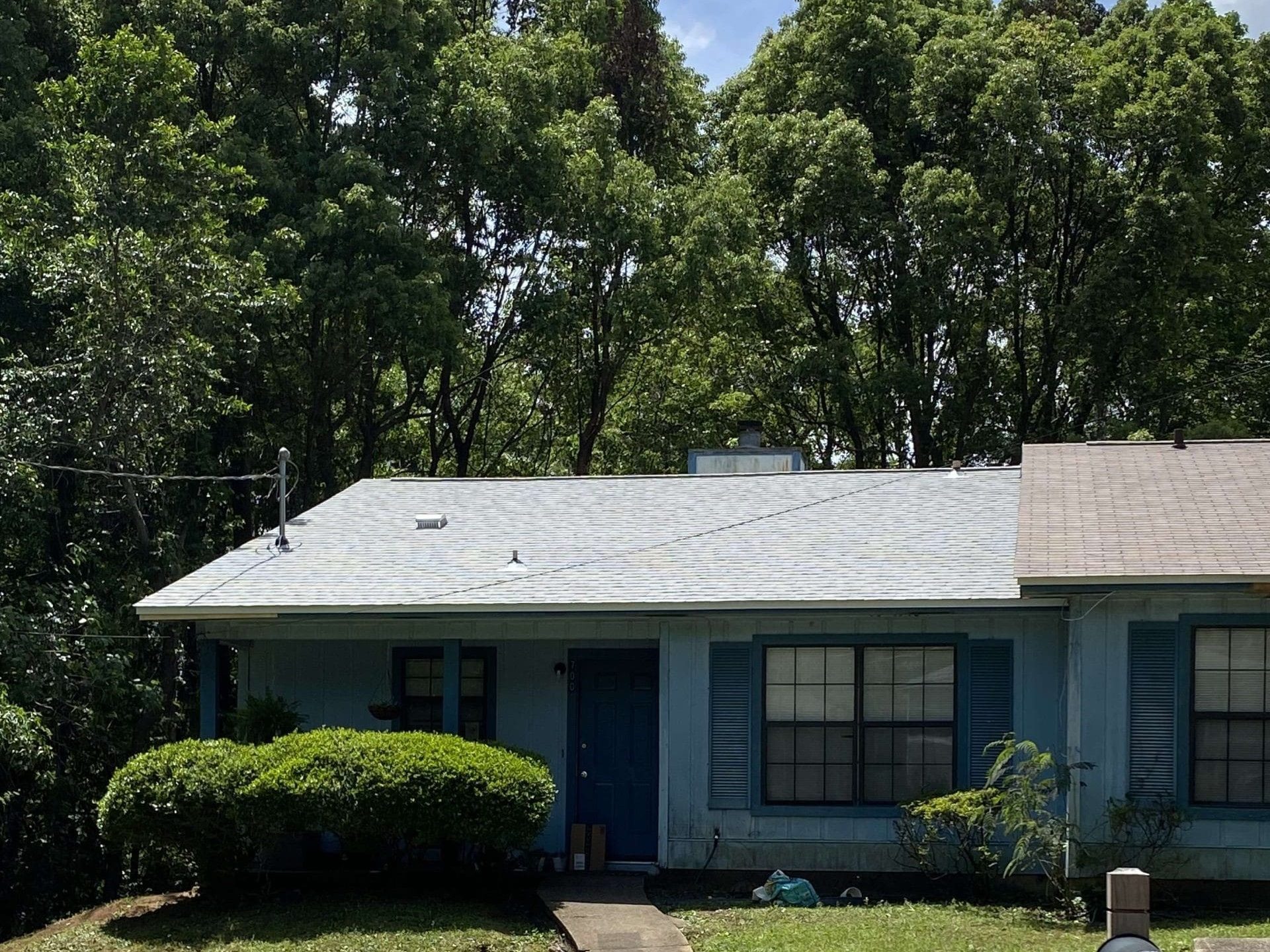 A light blue house with a grey roof, blue door, blue shutters, and trimmed bushes in front, set against a backdrop of tall green trees.