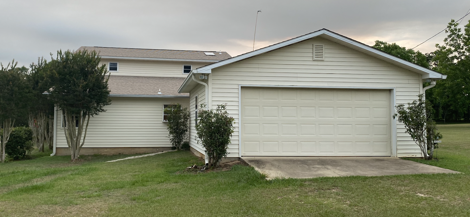 Single-story white garage with a closed white door, attached to a light-colored house, surrounded by green grass and shrubs under a cloudy sky.