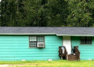 A small, single-story teal house with a dark roof, front steps, two windows, an air conditioning unit, and surrounded by green grass and trees.