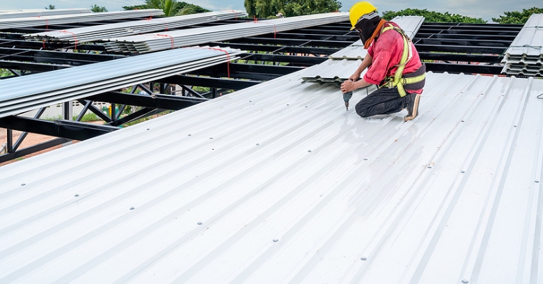 A construction worker in safety gear is installing metal roofing sheets on a building framework under an open sky.