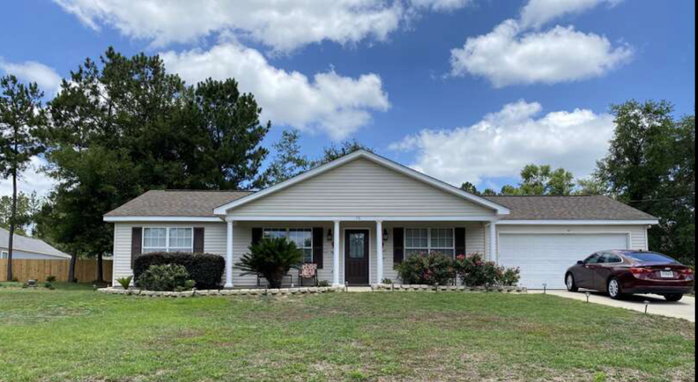 Single-story suburban house with a front lawn, attached garage, bushes along the walkway, trees in the background, and a red car parked in the driveway under a partly cloudy sky.