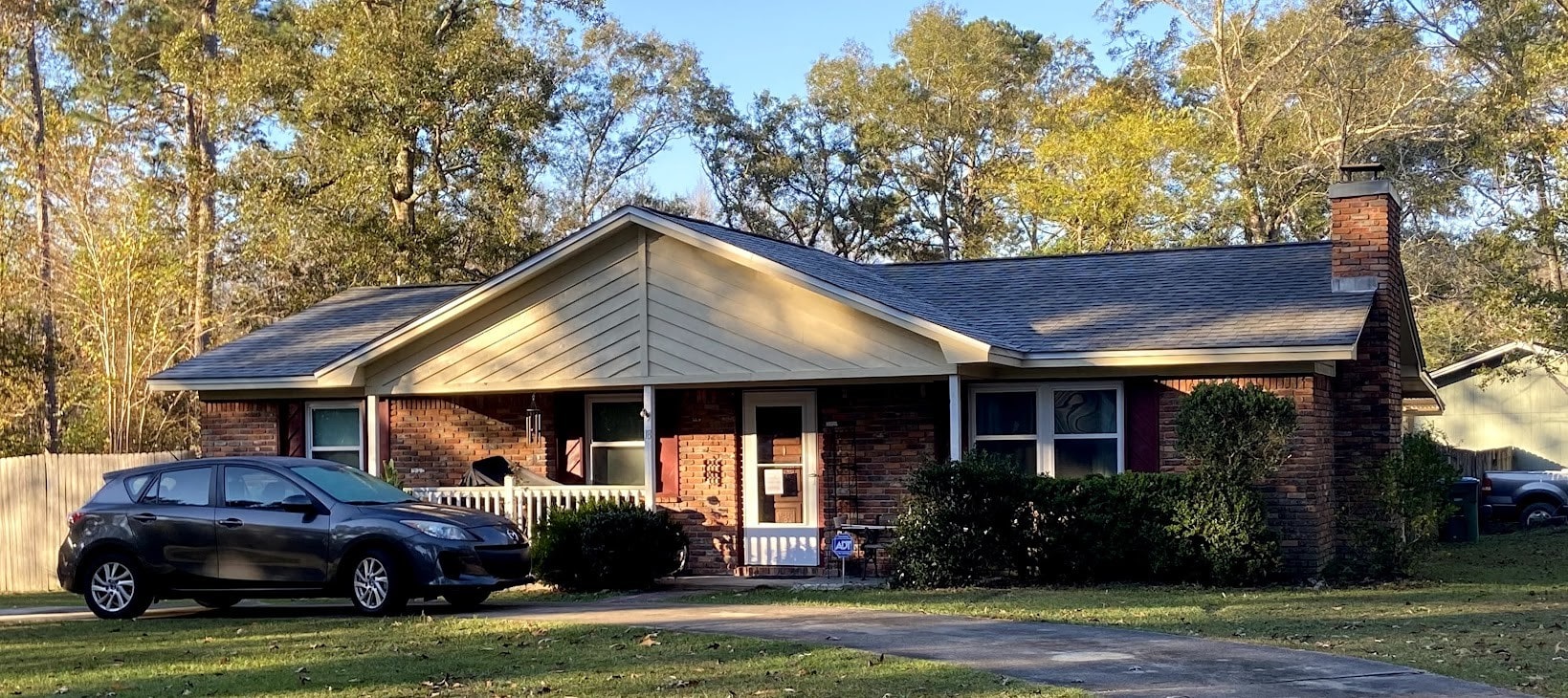 A single-story brick house with a covered porch, surrounded by bushes and trees, with a black car parked in the driveway on a sunny day.