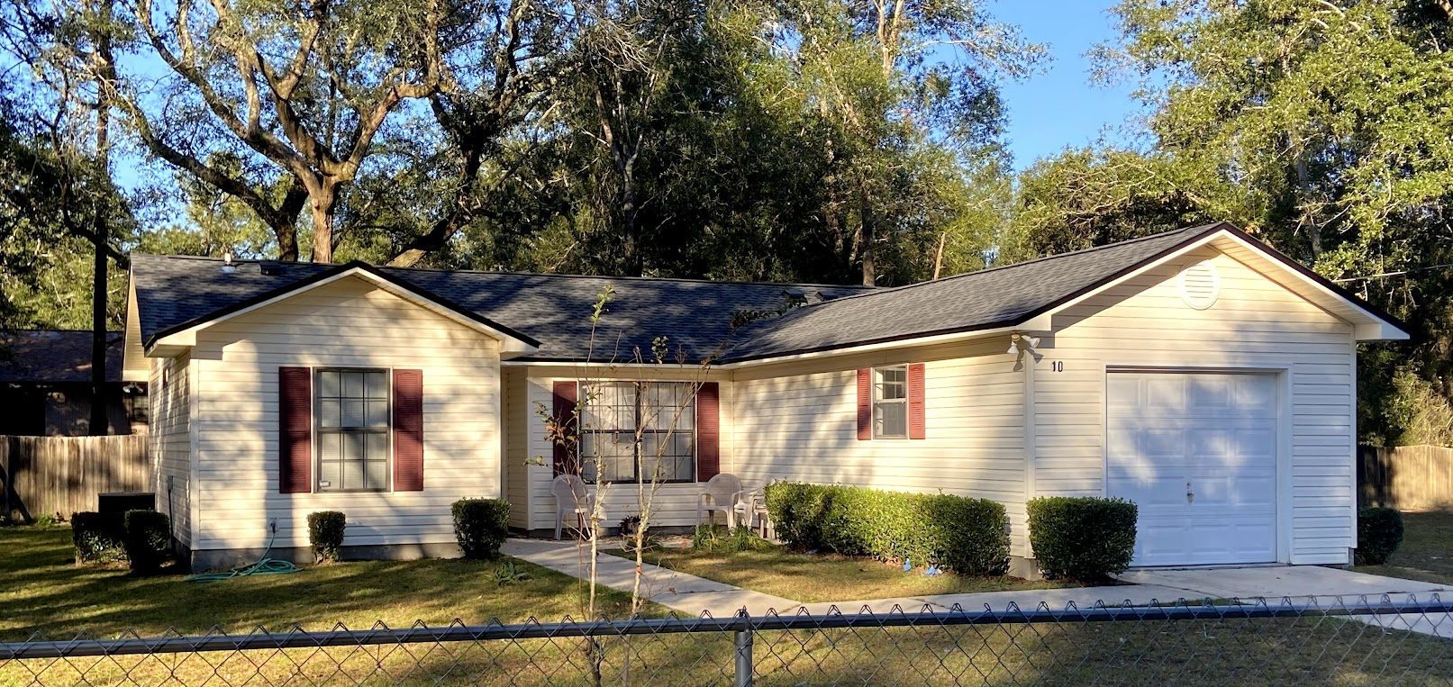 Single-story beige house with maroon shutters, a front porch, and an attached garage, surrounded by a lawn, bushes, and large trees, with a chain-link fence in front.