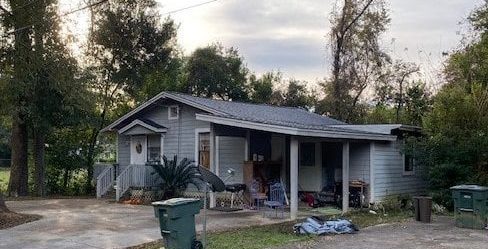 A small, gray house with a front porch, surrounded by trees, with trash bins and various items near the entrance.