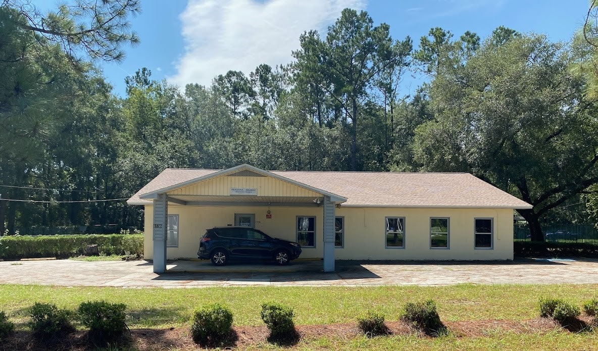 Single-story beige building with a covered entrance, one car parked under the awning, surrounded by trees and greenery on a sunny day.