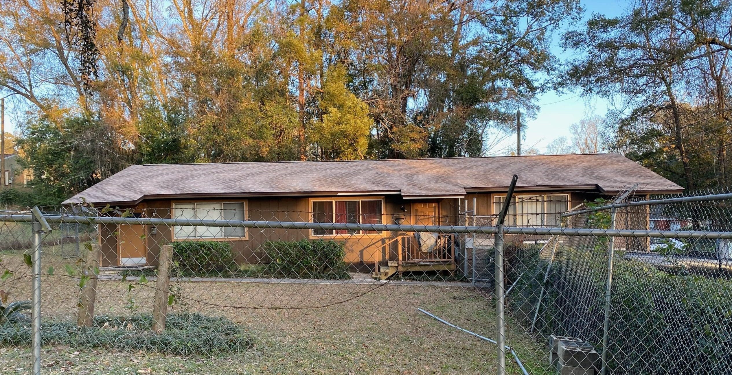 Single-story brown house with a front porch, behind a chain-link fence, and surrounded by trees in the background.