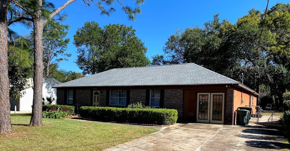 Single-story brick house with a gray roof, front lawn, driveway, and trees in the background under a clear blue sky.