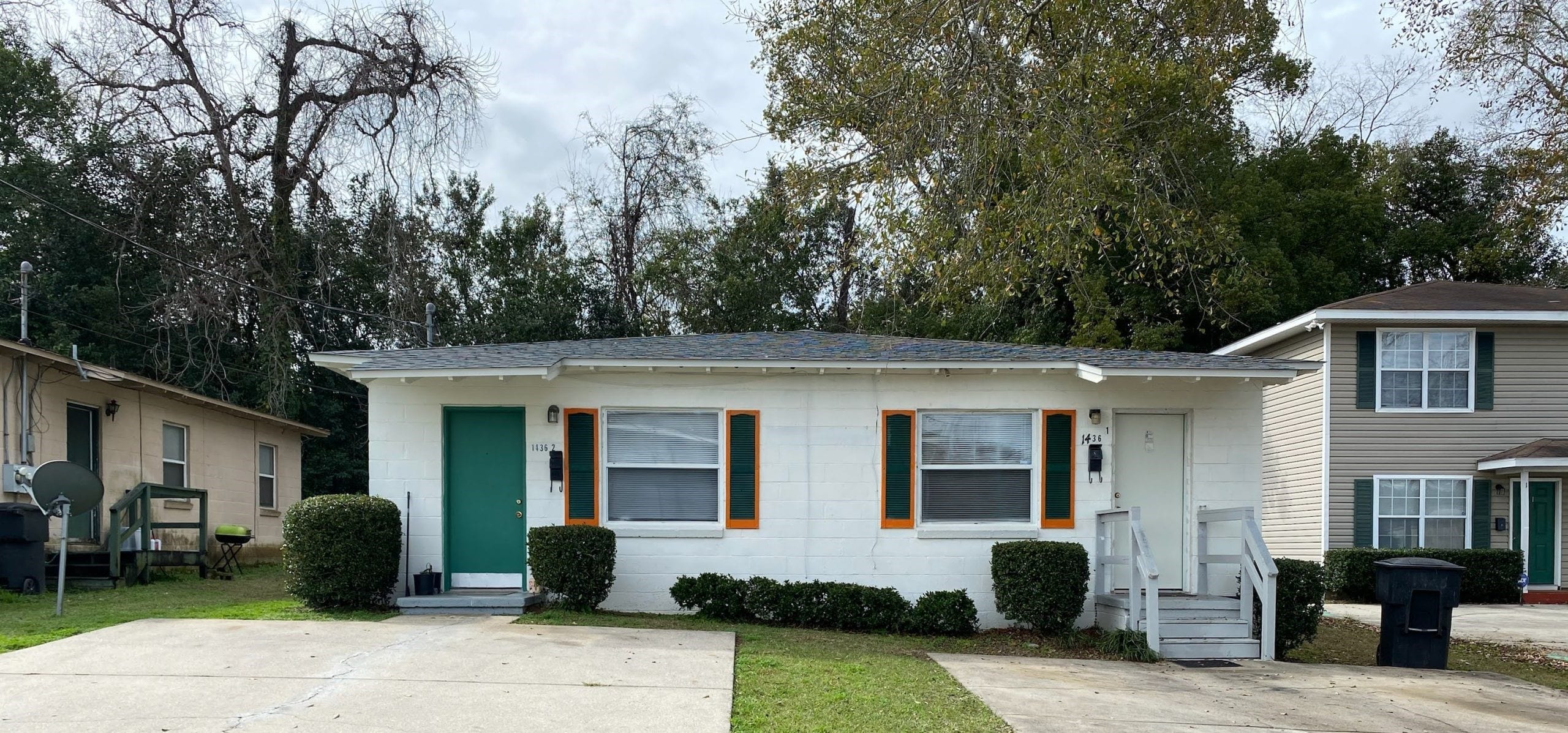 Single-story white house with orange-trimmed windows, green door, small front steps, bushes, and concrete driveway; trees and neighboring houses in the background.