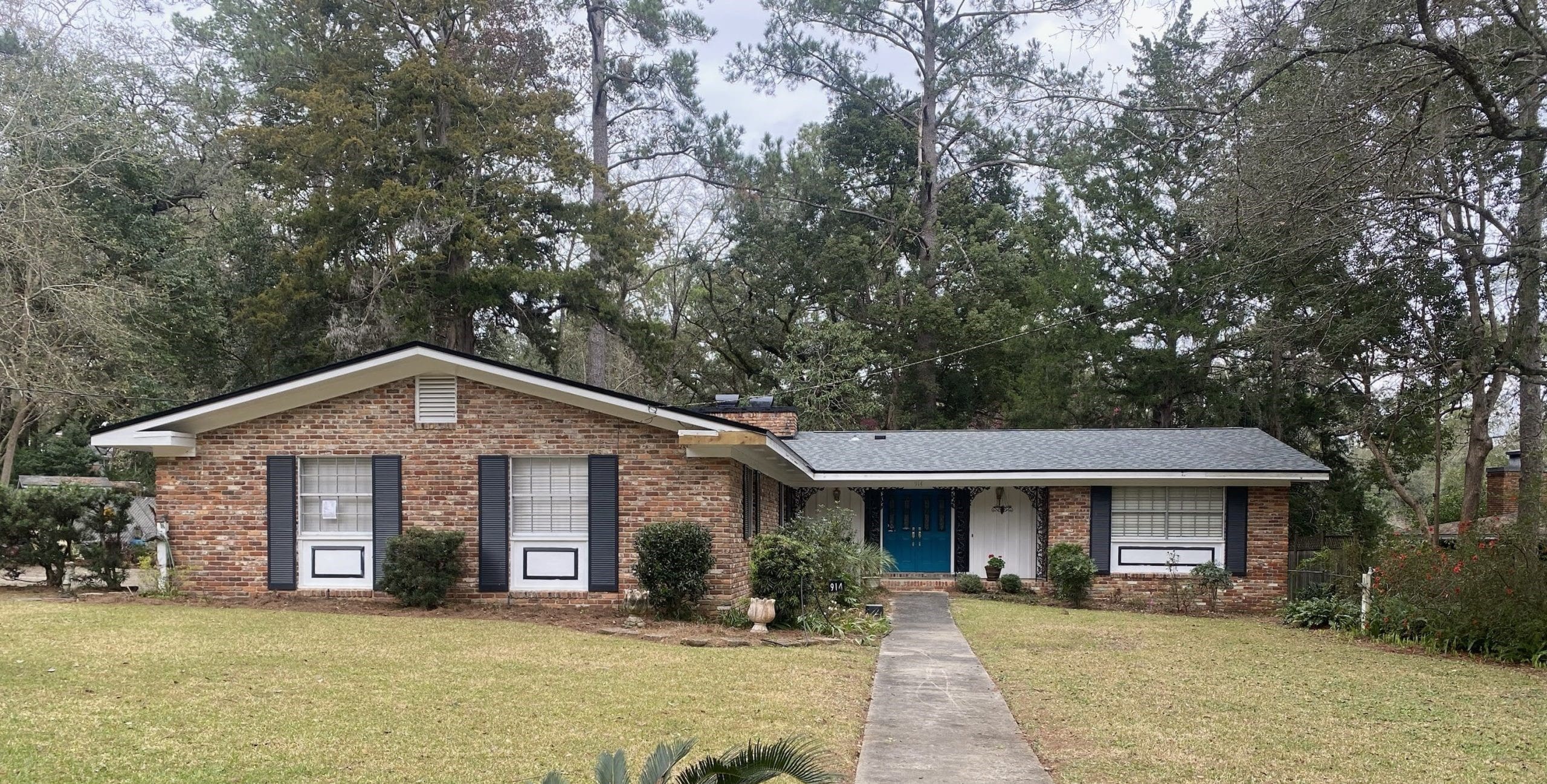 A single-story brick house with black shutters and a blue front door, surrounded by trees and a lawn with a concrete walkway leading to the entrance.