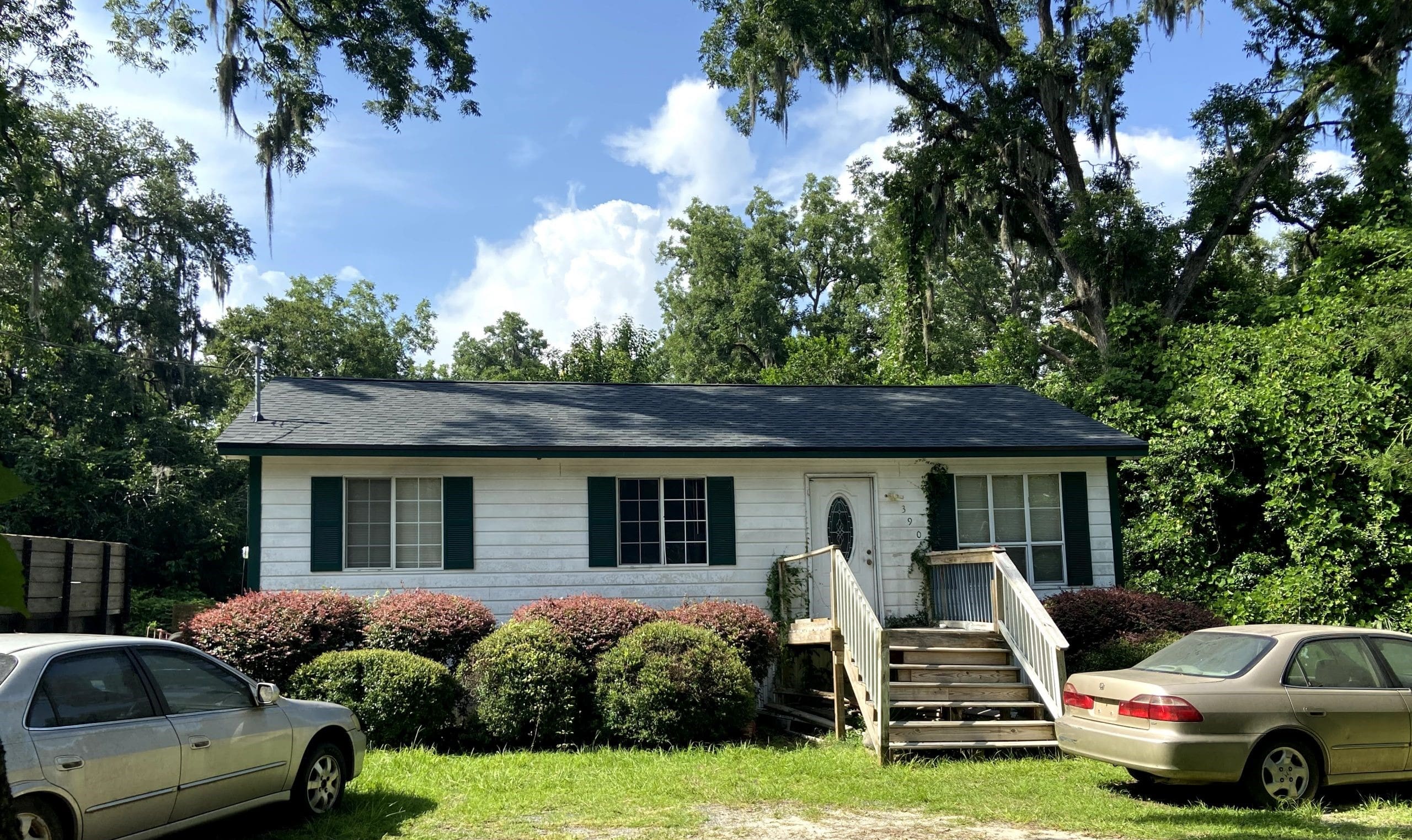 Single-story white house with dark shutters, front porch steps, and two parked cars in the yard, surrounded by trees and bushes under a partly cloudy sky.