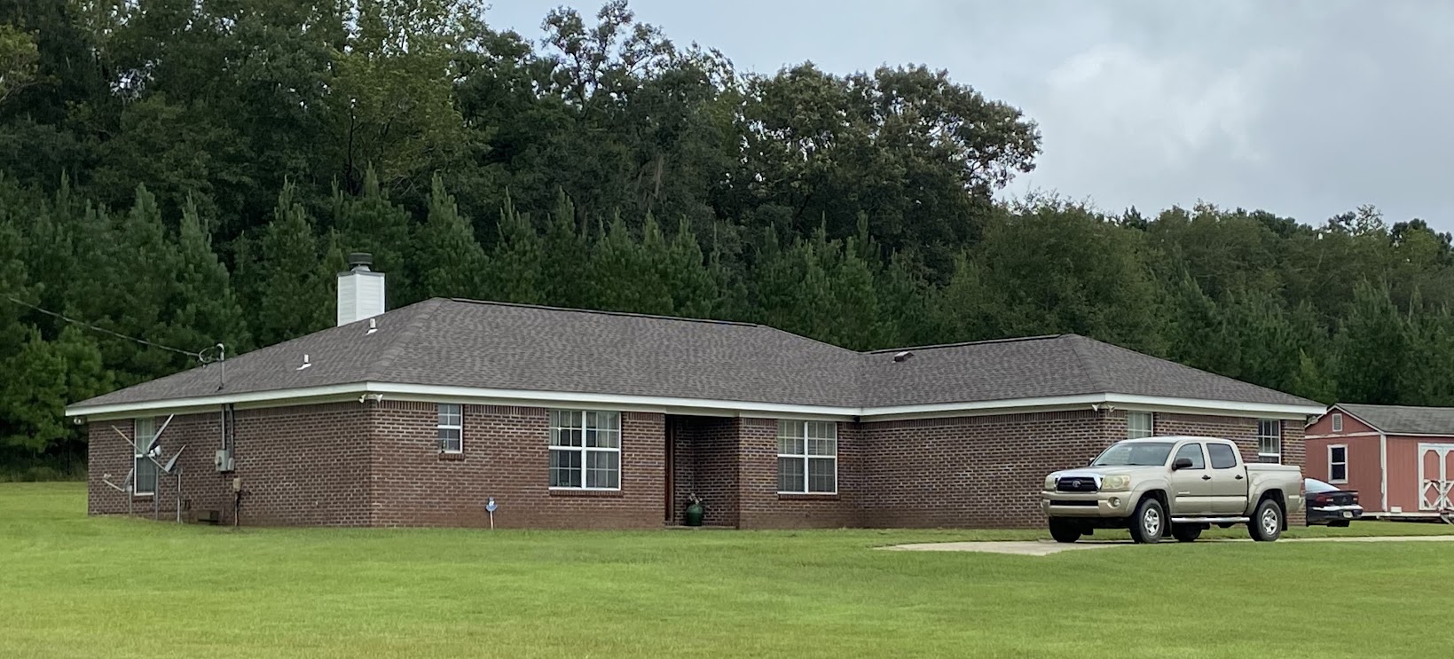 Single-story brick house with a gray roof, surrounded by a grassy yard, with a silver pickup truck parked in the driveway and a red shed in the background.