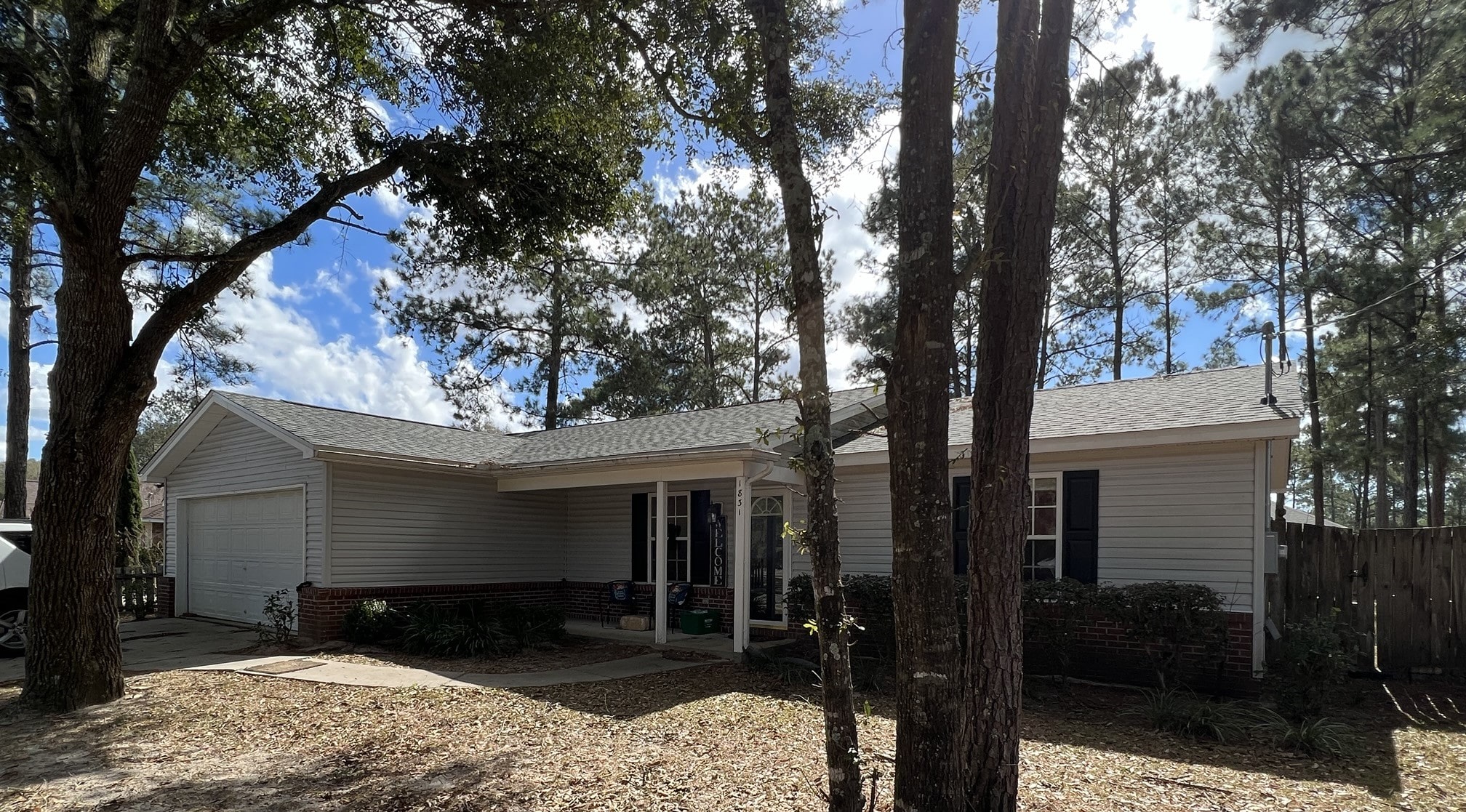 Single-story house with light-colored siding, brick accents, and a front porch, surrounded by tall trees under a partly cloudy sky.