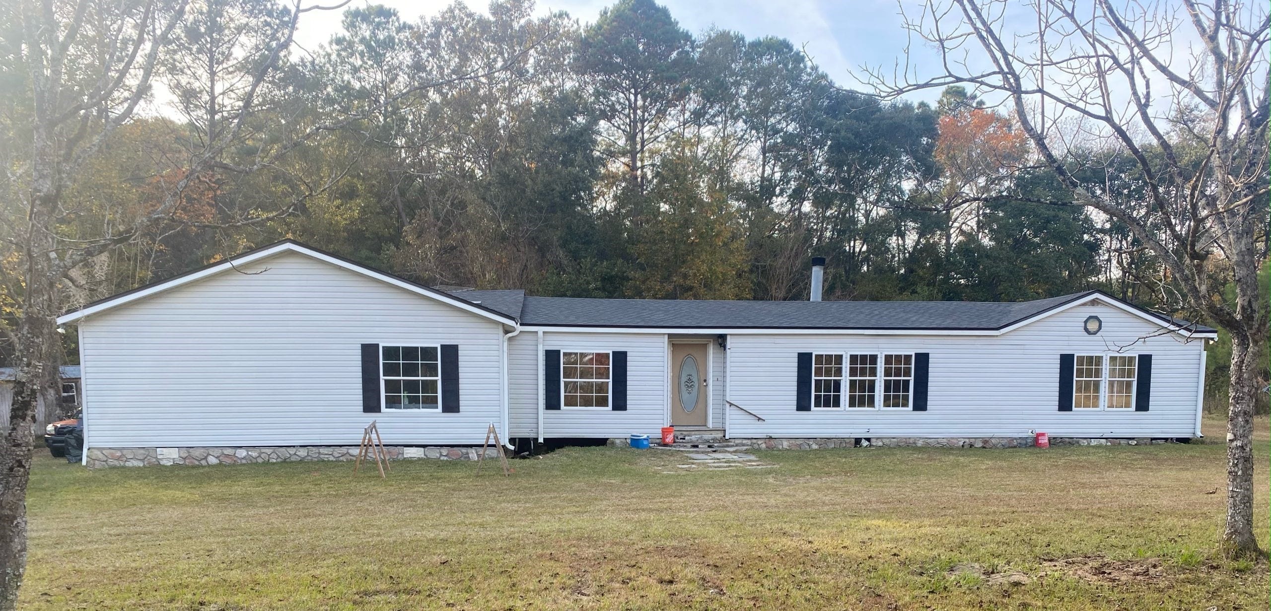 A single-story white manufactured home with black shutters sits on a grassy lawn, bordered by trees in the background.