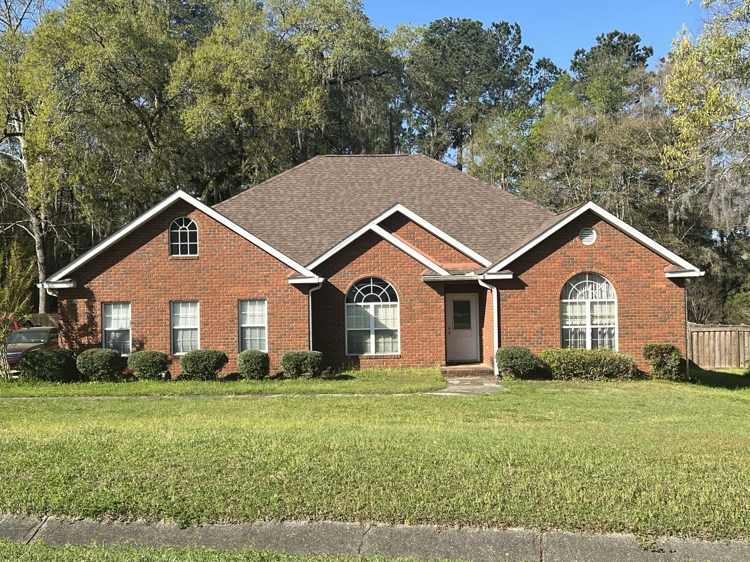 A single-story red brick house with a brown shingled roof, arched windows, and a neatly trimmed lawn in front. Trees are visible in the background.