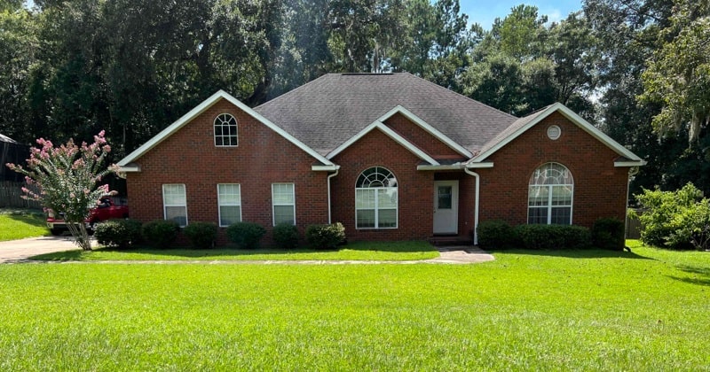 Single-story red brick house with a dark roof, several windows, and a front yard with green grass and shrubs, set against a backdrop of tall trees. Single-story red brick house with a dark roof, several windows, and a front yard with green grass and shrubs, set against a backdrop of tall trees.