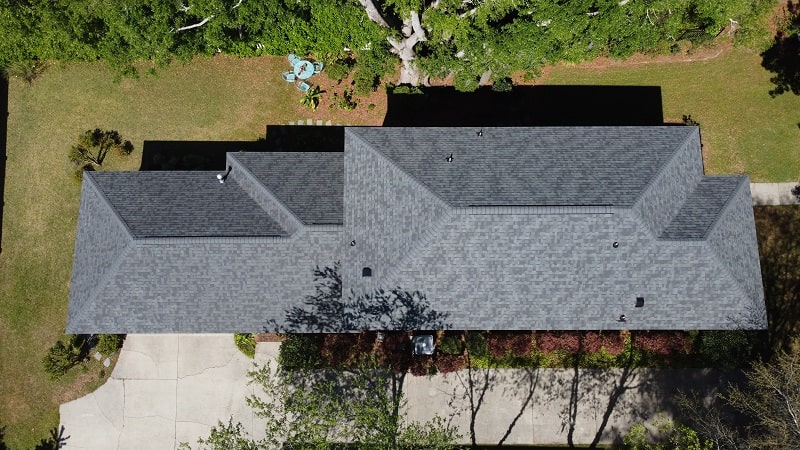 Aerial view of a gray shingle roof on a single-story house, surrounded by trees, lawn, driveway, and landscaping. Aerial view of a gray shingle roof on a single-story house, surrounded by trees, lawn, driveway, and landscaping.