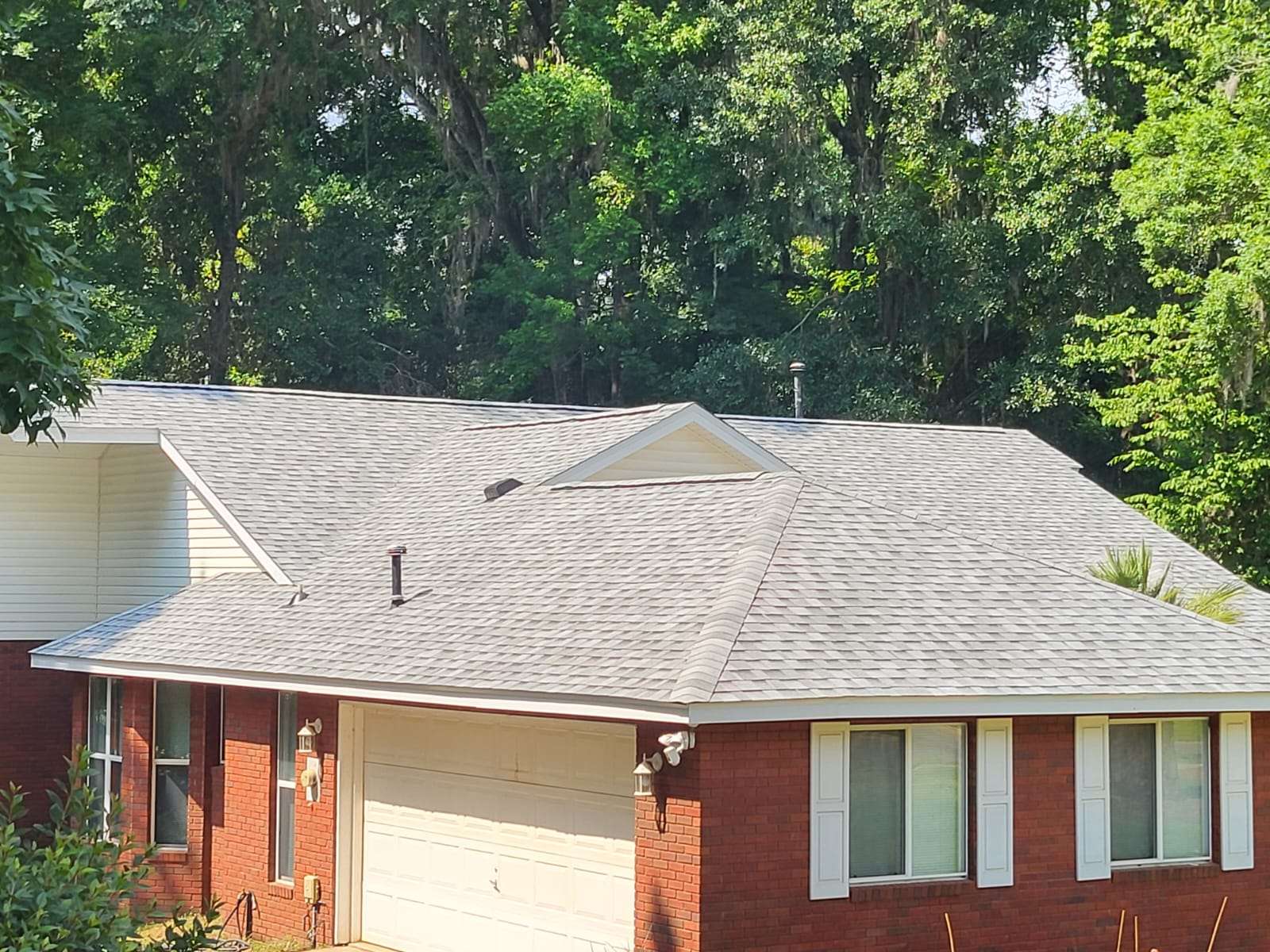 A brick house with white trim and a light gray shingle roof, surrounded by tall green trees in the background.