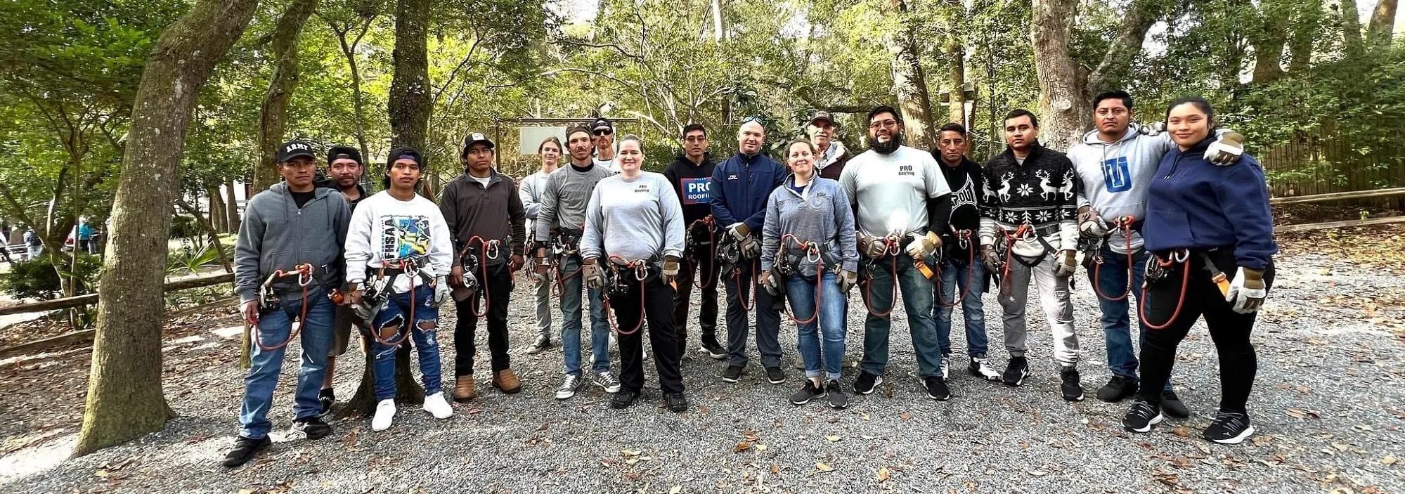 A group of people wearing harnesses and gloves stand in a wooded outdoor area, posing together on a gravel surface.