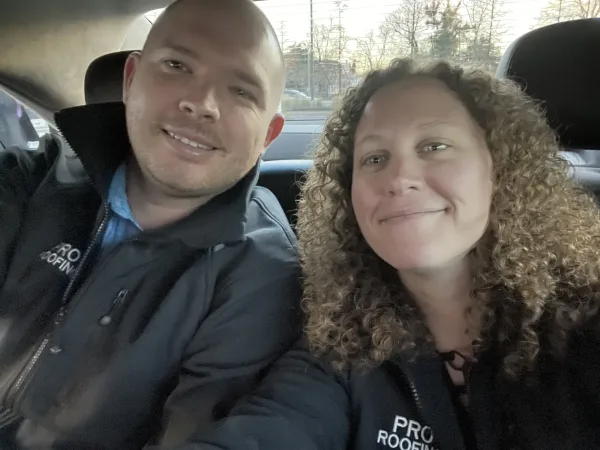 A man and a woman sit side by side in a car, both smiling at the camera and wearing jackets with PRO ROOFING logos.