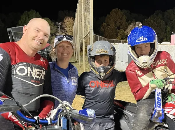 Four people pose together at an outdoor dirt track at night; two are wearing motocross gear and helmets, holding motorcycles.