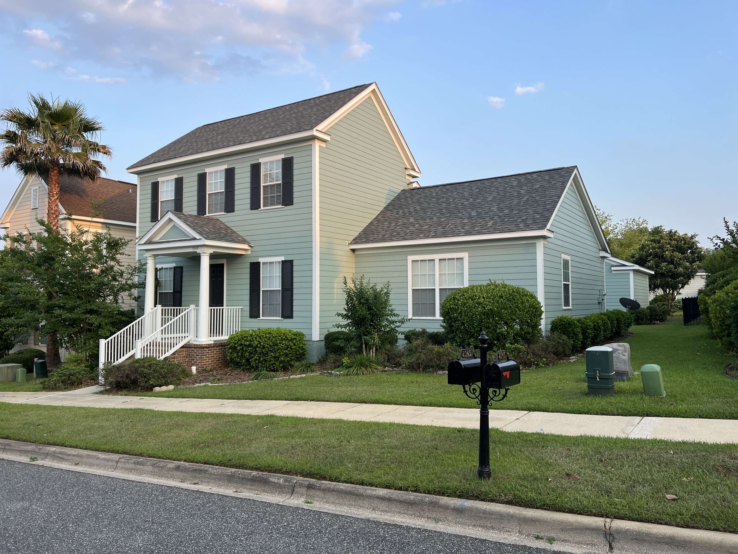 Light green two-story house with white trim, black shutters, and a front porch. Lawn, bushes, mailbox, and utility boxes in front. Sidewalk runs along the street.