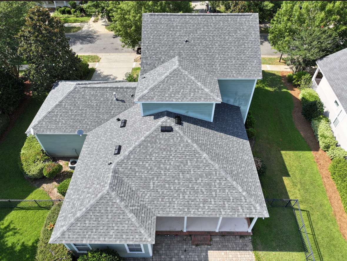 Aerial view of a light blue house with grey shingle roof, attached garage, green lawns, trees, and a paved walkway leading to the entrance.