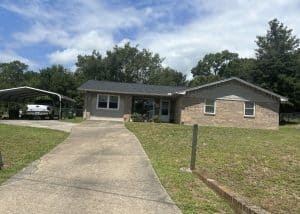 Single-story brick house with a black roof, wide driveway, carport on the left with a white vehicle, and a large front yard with minimal landscaping.